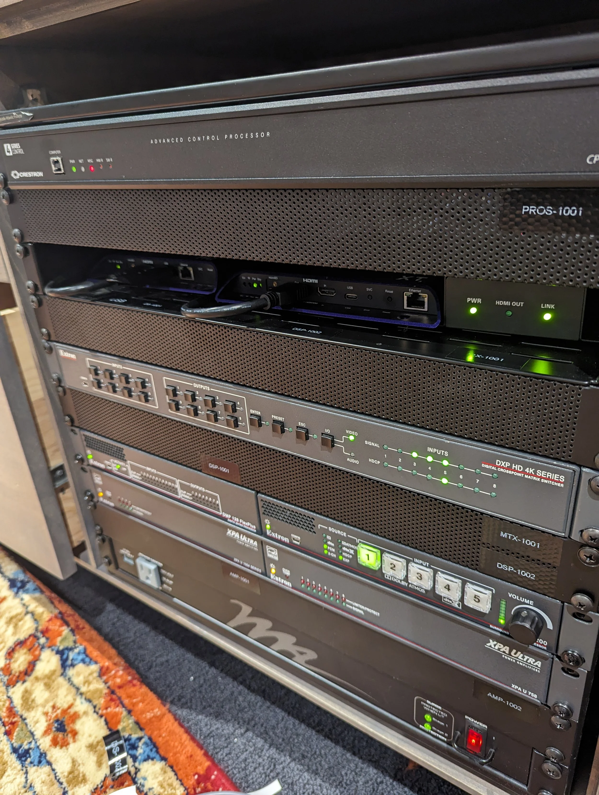 Audio-visual equipment rack with various electronic devices including a control processor, switcher, and amplifier, installed in a room with a carpeted floor.