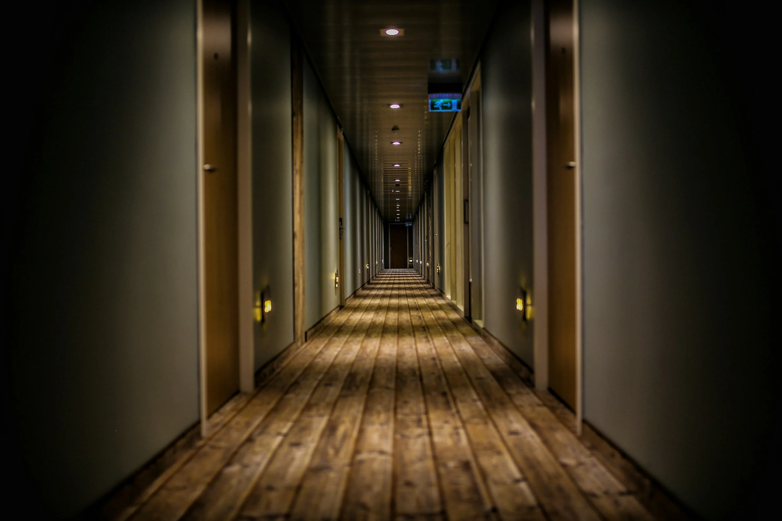 Long hotel hallway with wood flooring, closed doors on both sides, and ceiling lights. There are signs and lights at the end of the corridor.