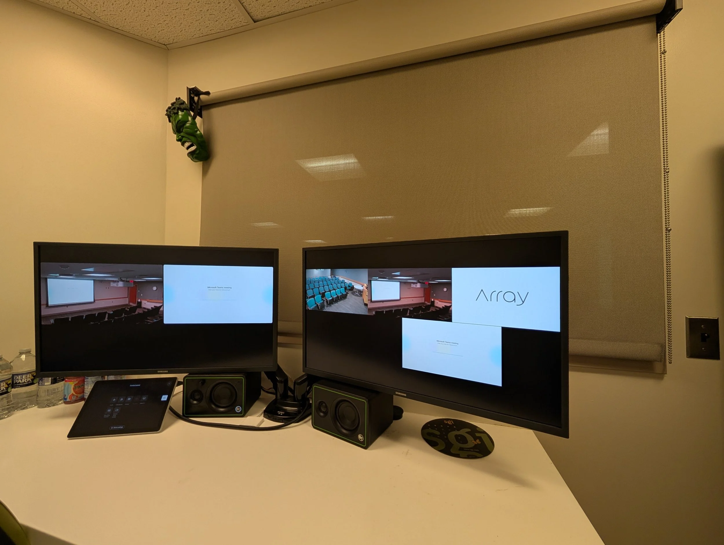 Two computer monitors on a white desk displaying a presentation on a classroom or conference room with a projector and empty chairs. There are two speakers, a small control panel, and some water bottles on the desk. A beige wall with a white roller s