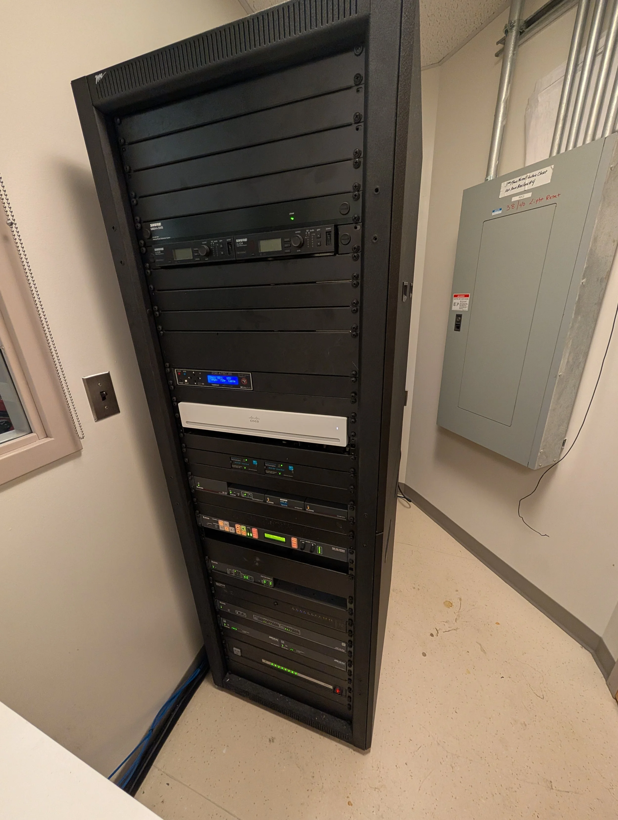 A black server rack filled with networking equipment against a beige wall, with a white air conditioning unit on the wall nearby.