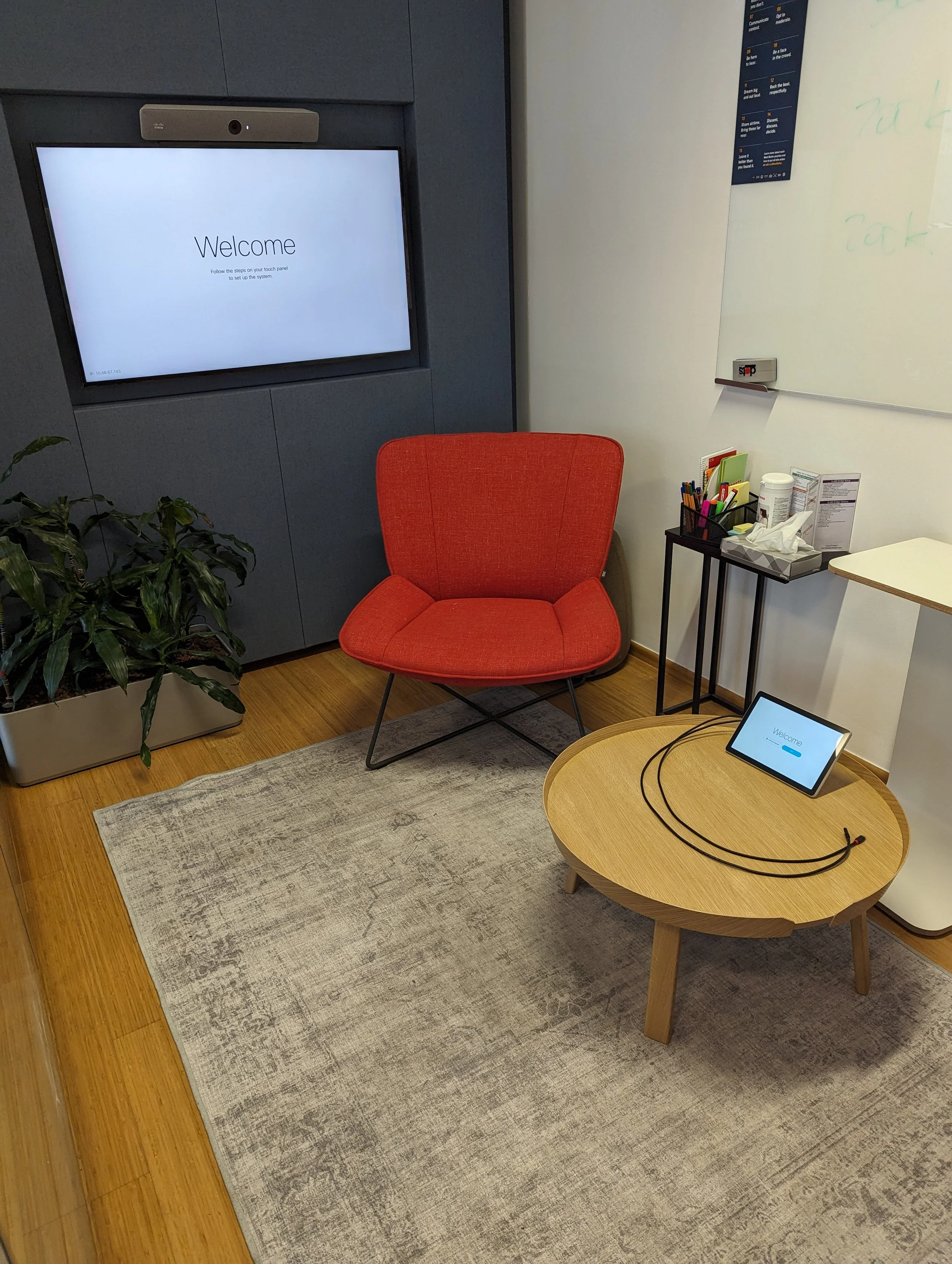 An office waiting area with a red chair, a small round wooden table with a tablet on it, a whiteboard, and a large screen displaying a welcome message.