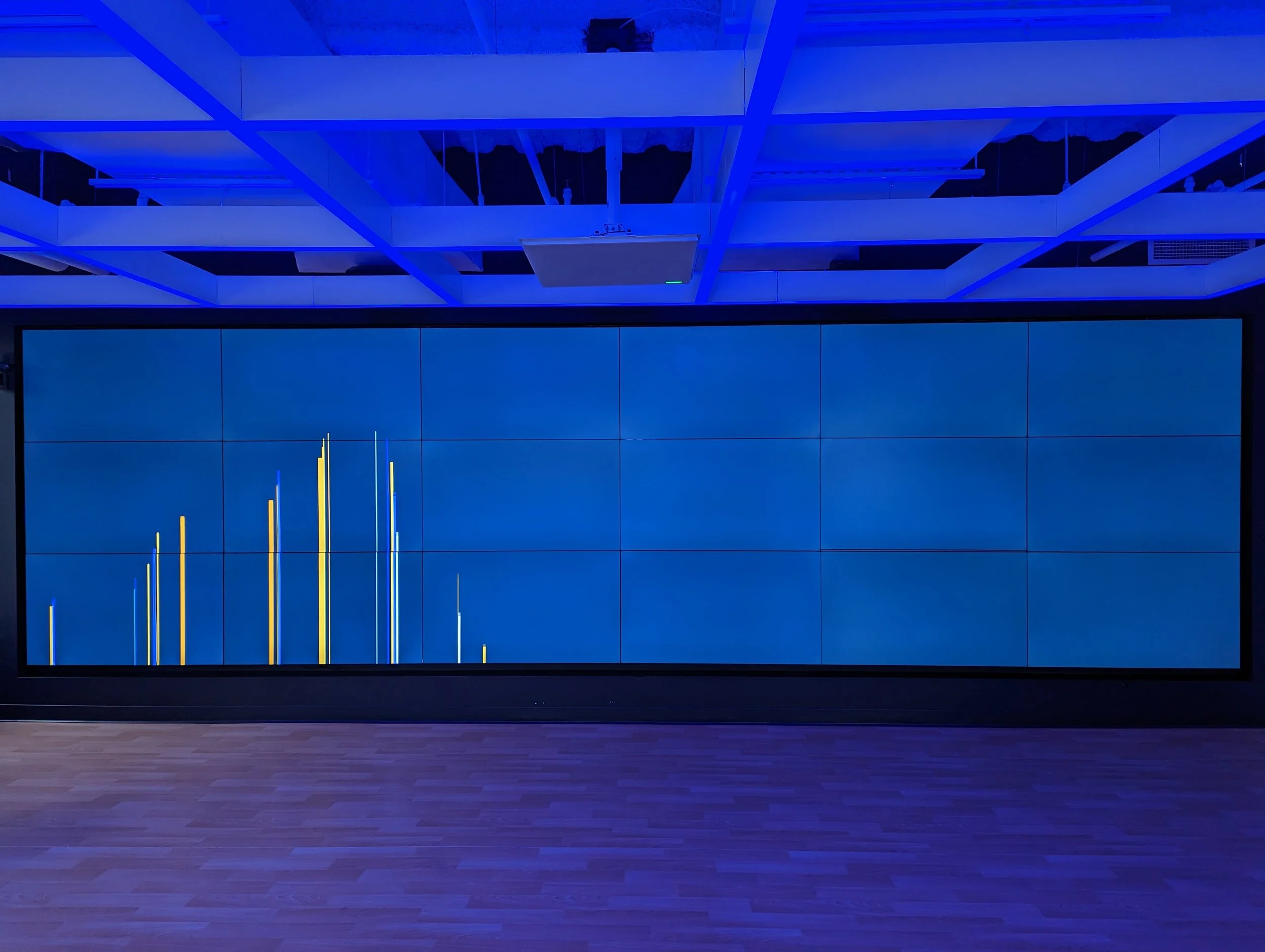 Large digital display wall showing a blue background with yellow and blue bar graphs. The room has a wooden floor and ceiling with rectangular lighting panels.