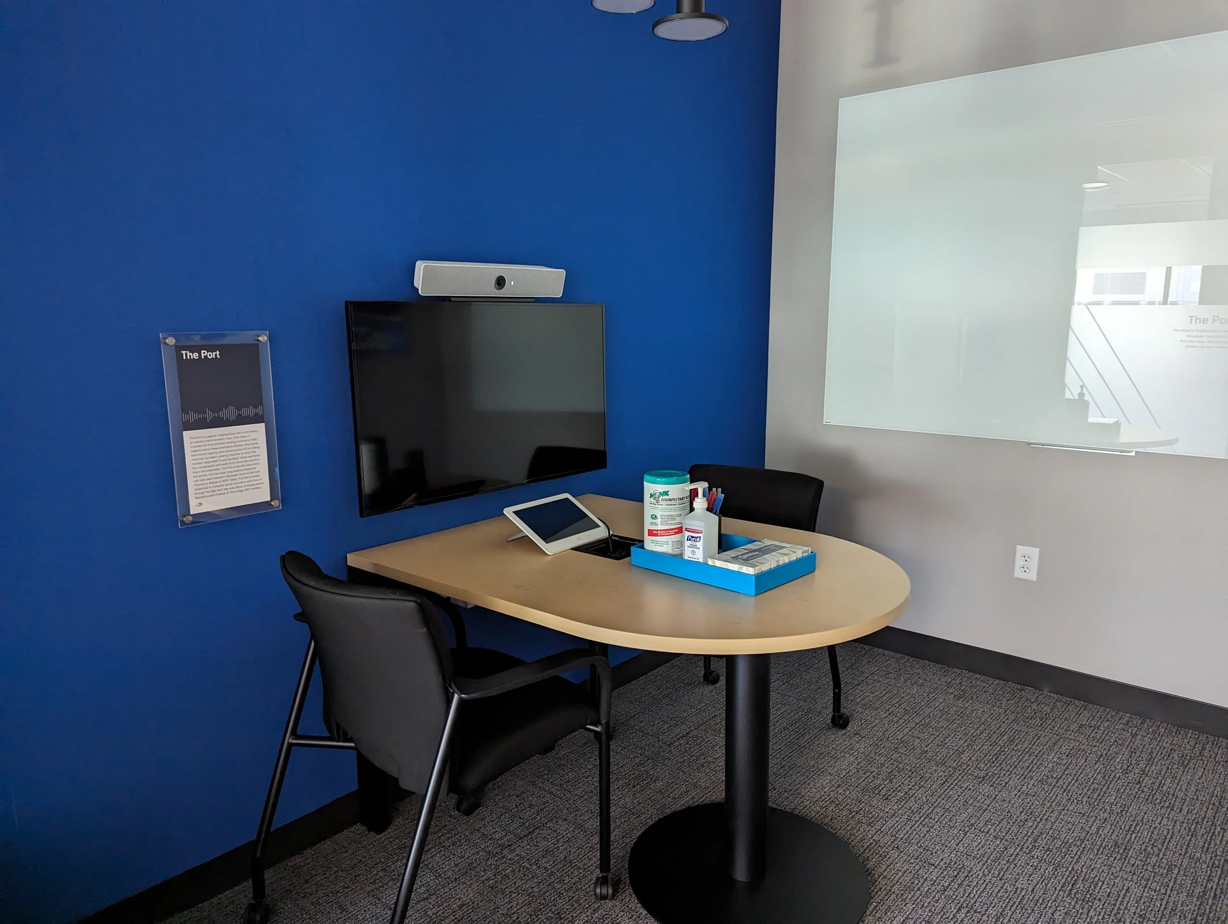 An office or meeting room with a wooden table, black chairs, a wall-mounted monitor, a screen, and supplies on the table, with a blue wall and white wall with a glass reflection.