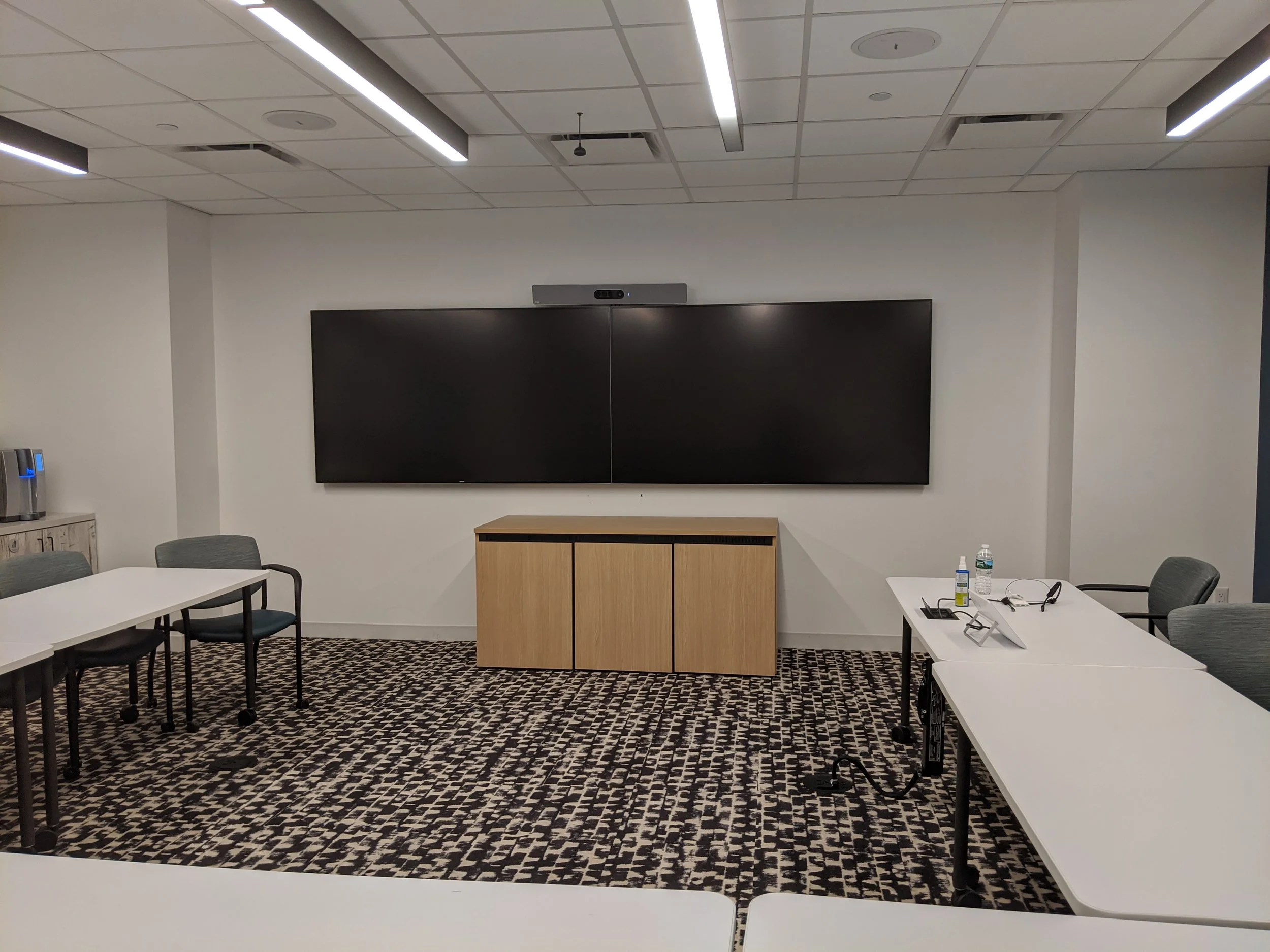 Empty conference room with a large digital screen on the wall, a wooden cabinet below it, and white tables with chairs arranged around. Items on the tables include a water bottle, sanitizer, and electronic devices.