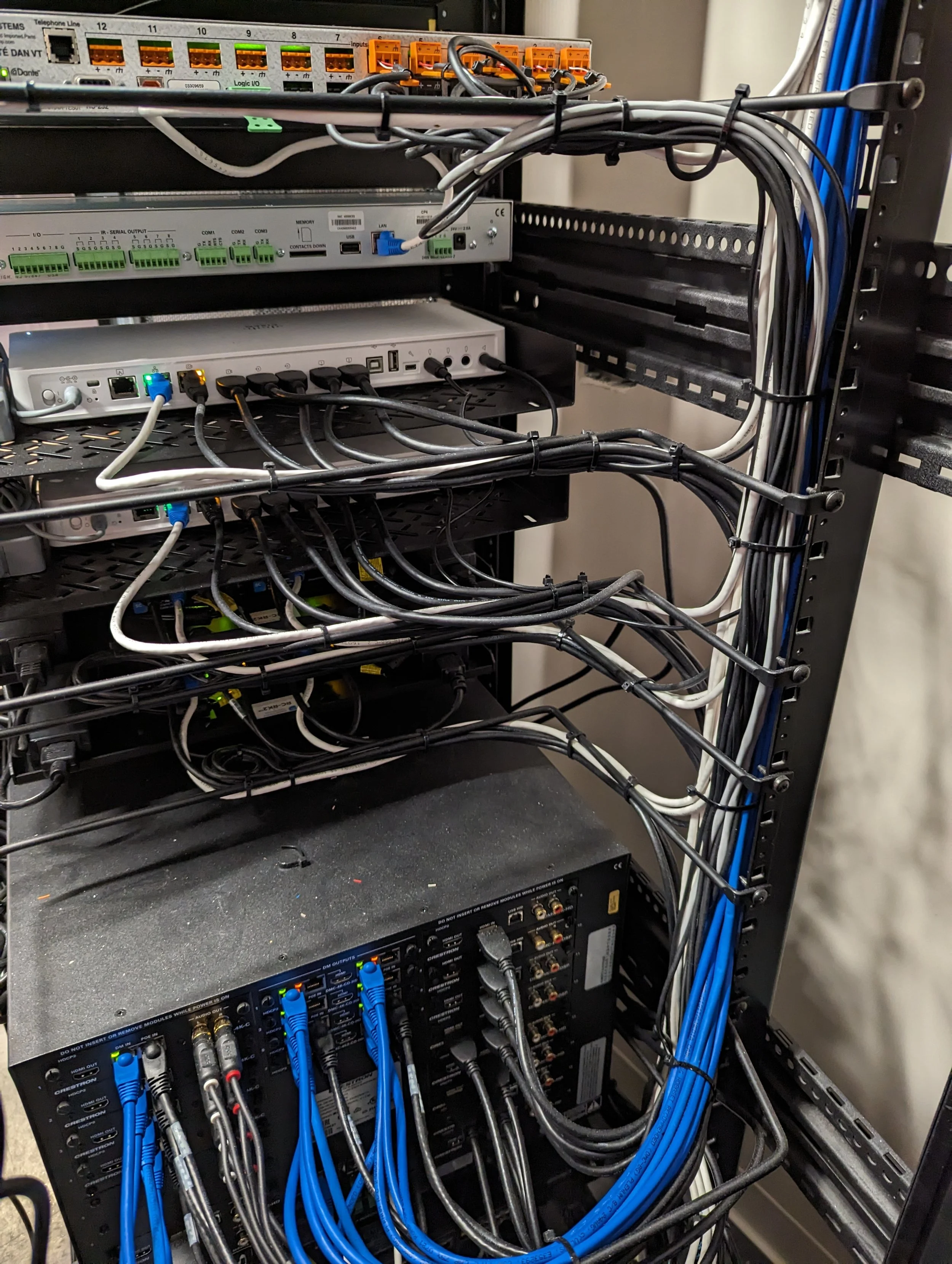 A network server rack containing various electronic equipment, including switches, routers, and patch panels, with tangled black and blue Ethernet cables connected to the devices.