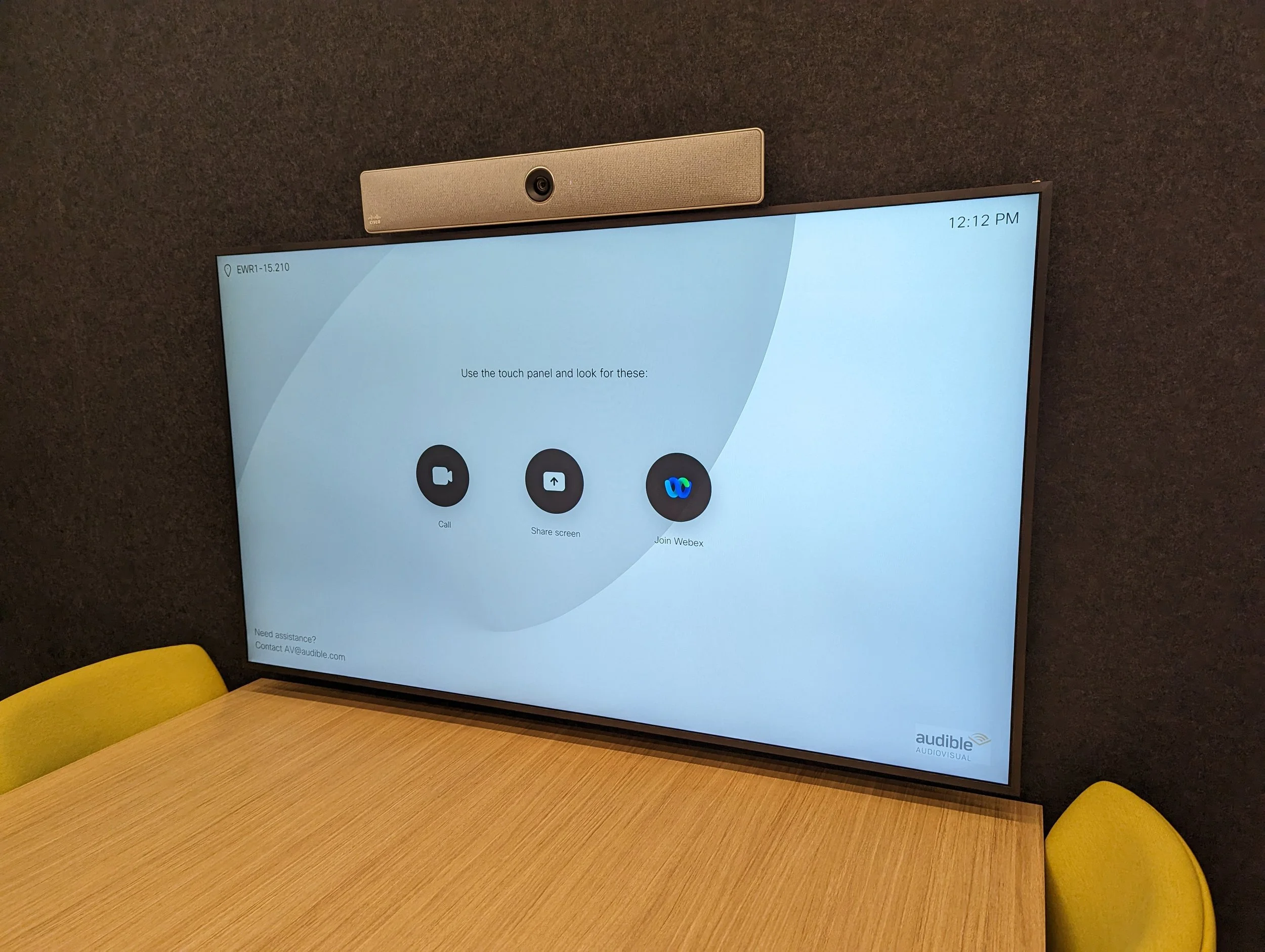 A large conference room screen displaying Webex meeting options, mounted above a wooden table with yellow chairs.
