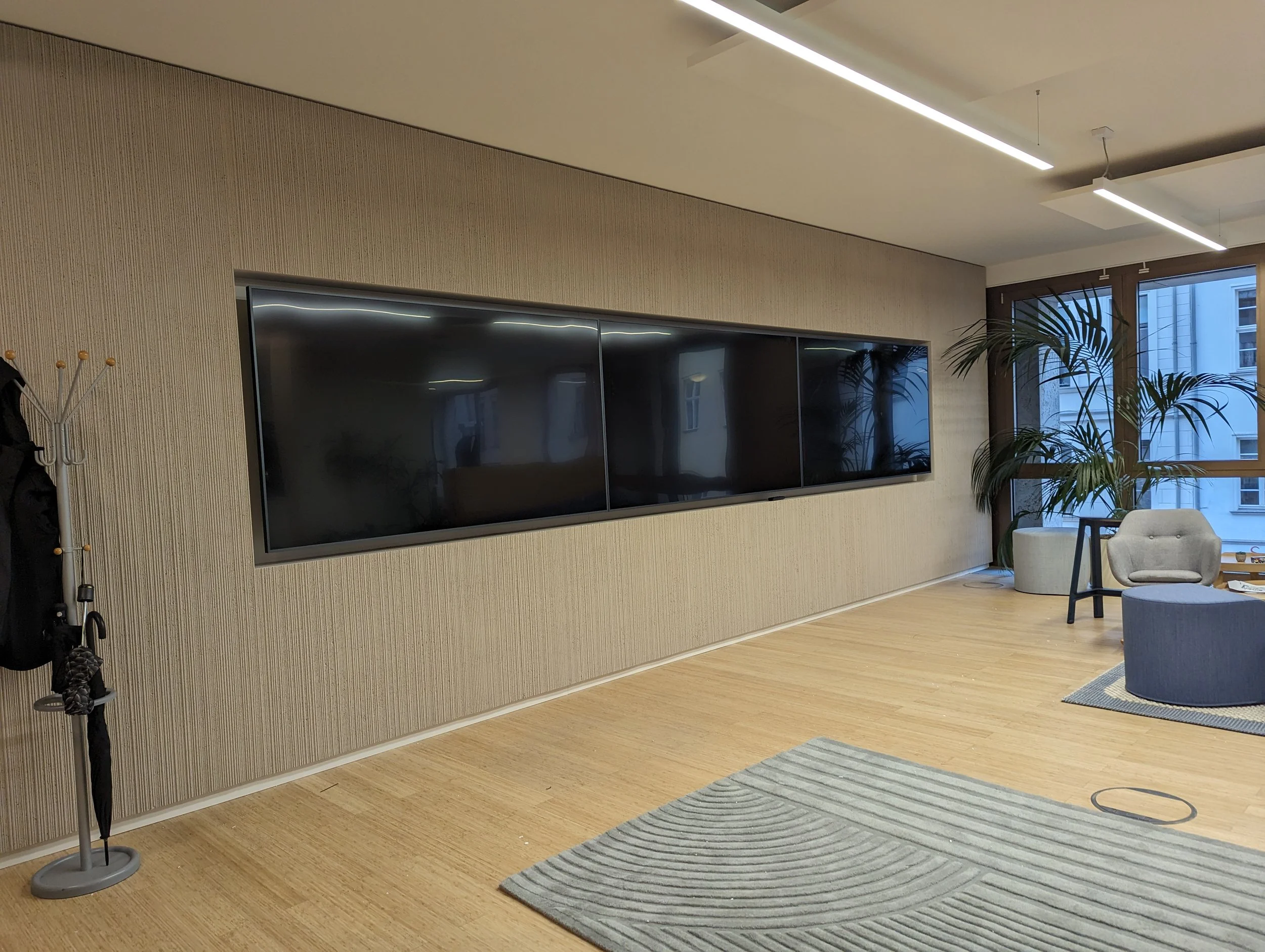 Modern living room with three large black flat-screen TVs mounted on a textured beige wall, a potted plant near a window, and a cozy sitting area with a light-colored chair and a small round ottoman, with a door and city buildings visible through the