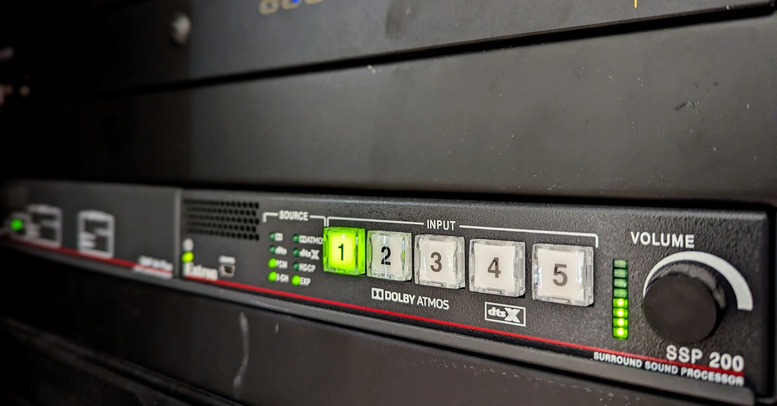 Close-up of a black surround sound receiver with illuminated input and volume control buttons, labeled 'SSR 200,' featuring LED indicators and a volume knob.