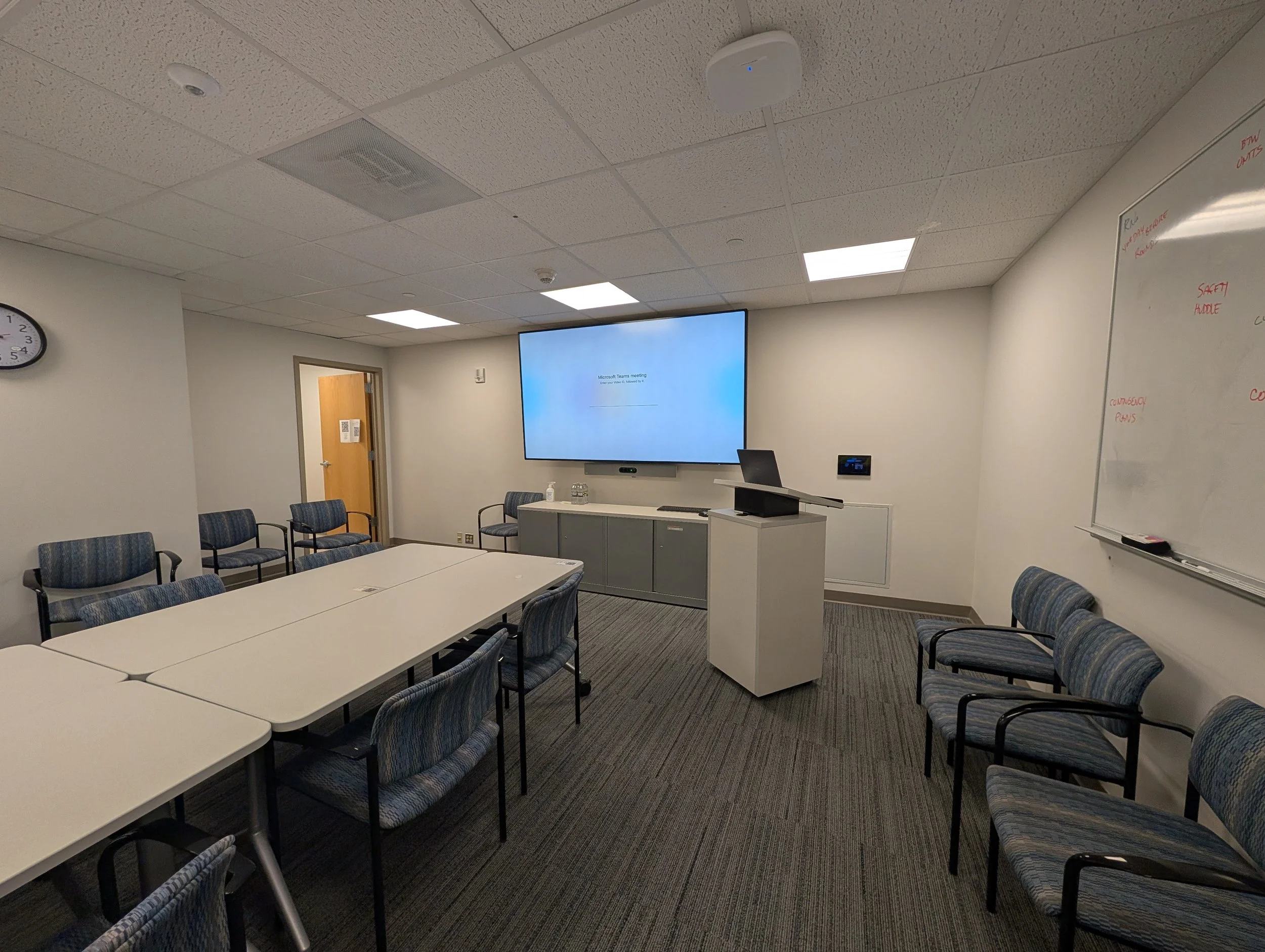 Empty corporate conference room with white tables arranged in a U-shape, blue striped chairs, a large screen displaying a Windows login screen, a whiteboard with red writing, and a clock showing 4:37.