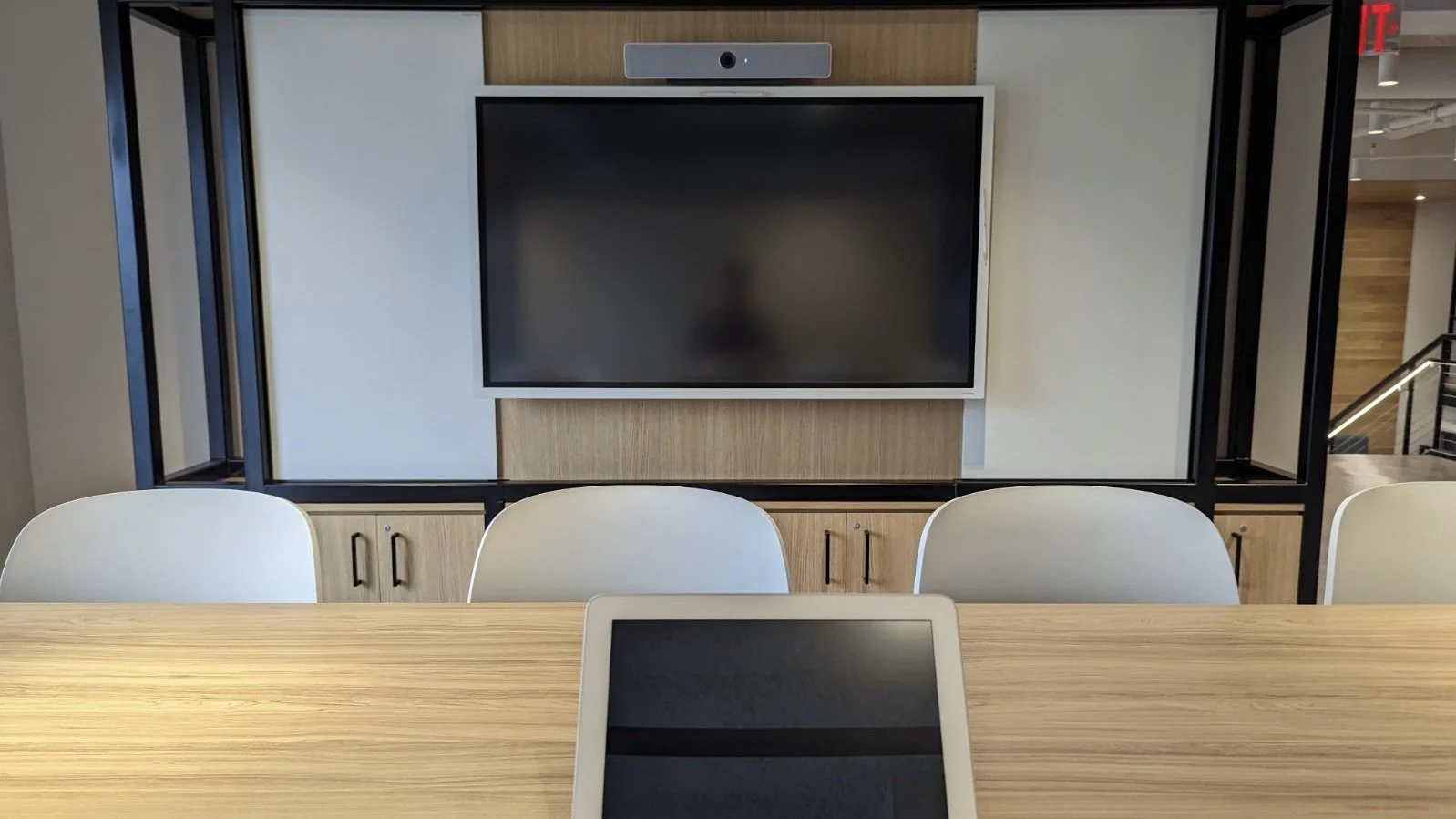 Conference room with a wooden table, three white chairs, a large black screen mounted on the wall, and a video conferencing camera above the screen in a modern office space.