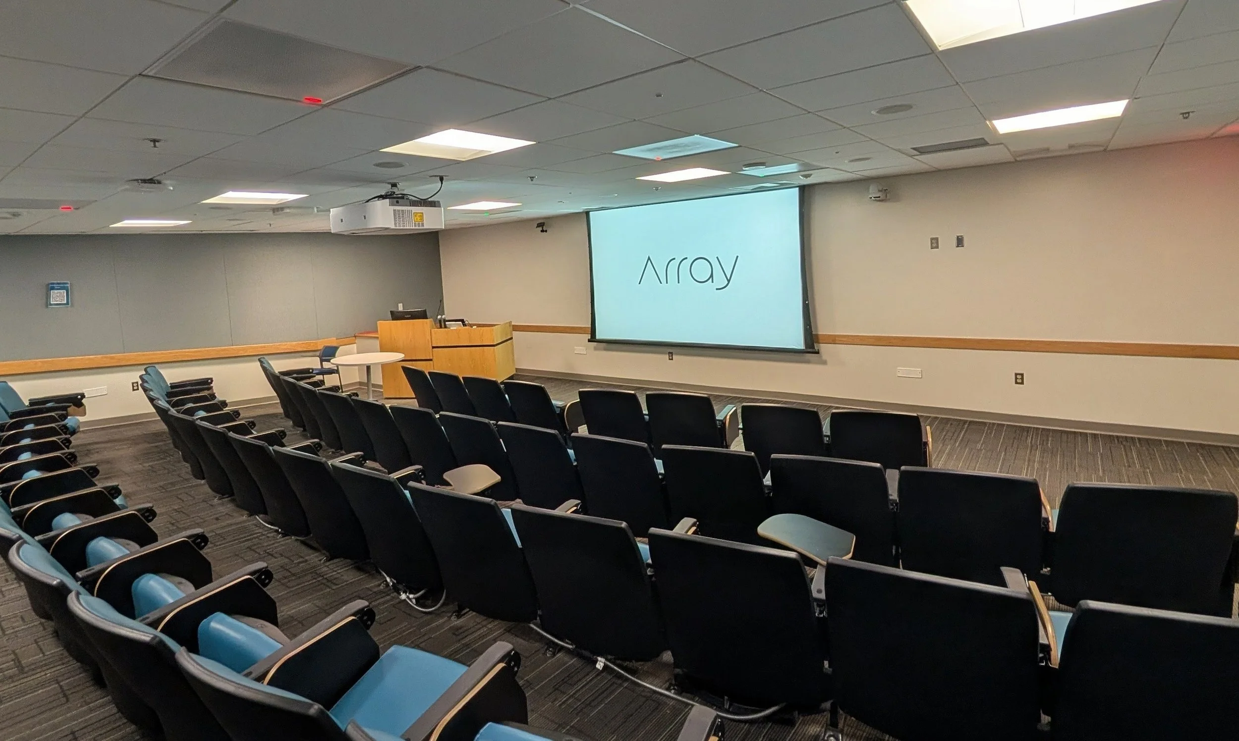 Empty conference room with black chairs, a projector screen displaying the word 'Array', a podium, and ceiling lights.