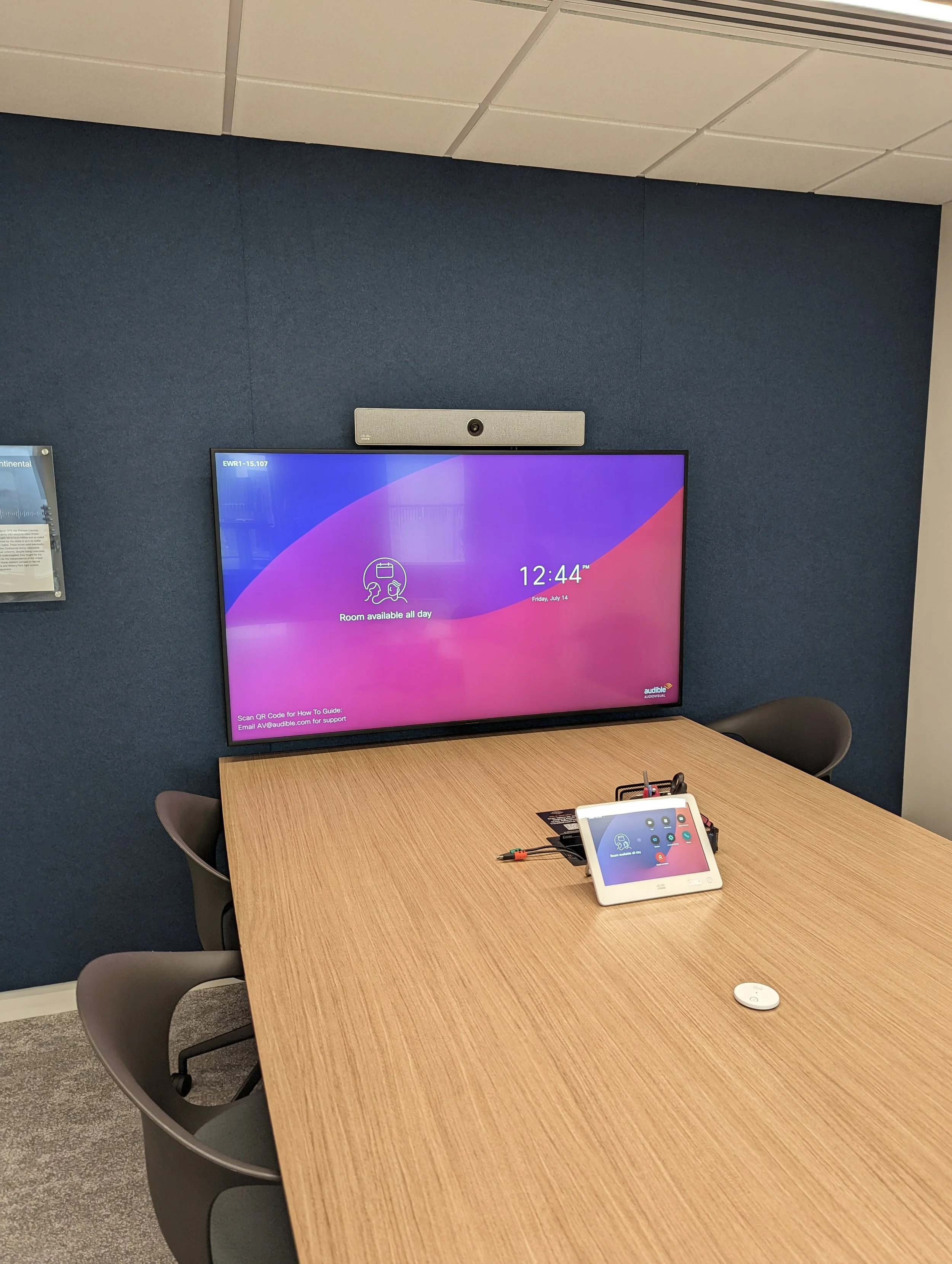 A conference room with a large wall-mounted screen displaying the time, date, and a message that says the room is available all day, with a small tablet on the table showing meeting options and a white device on the table.