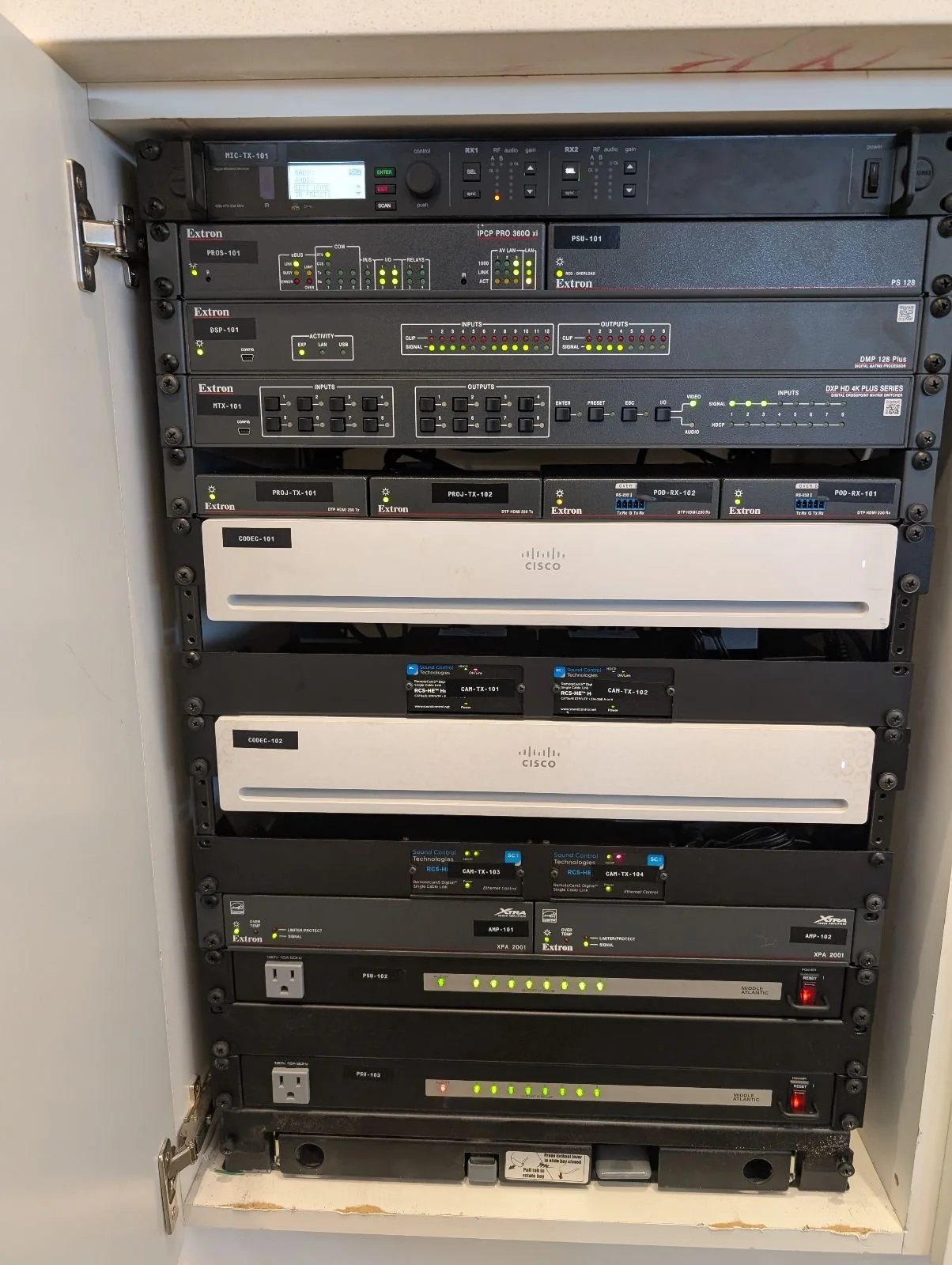 Server rack containing networking and audio-visual equipment including Cisco devices, Extron controllers, and power strips.