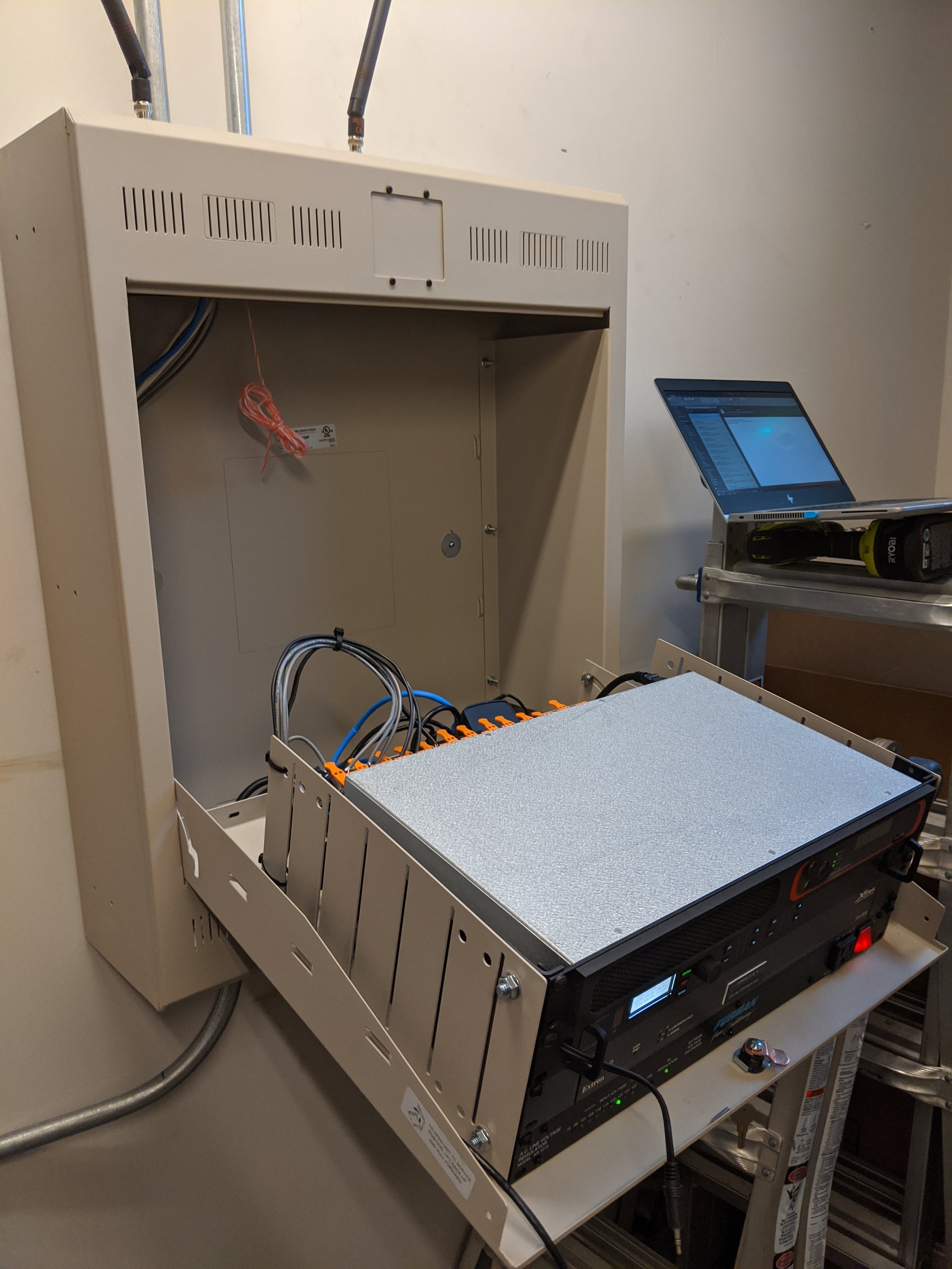 An open electrical or network equipment cabinet on a table with electronic testing or server equipment connected, a laptop on a nearby stand, and wires inside the cabinet.