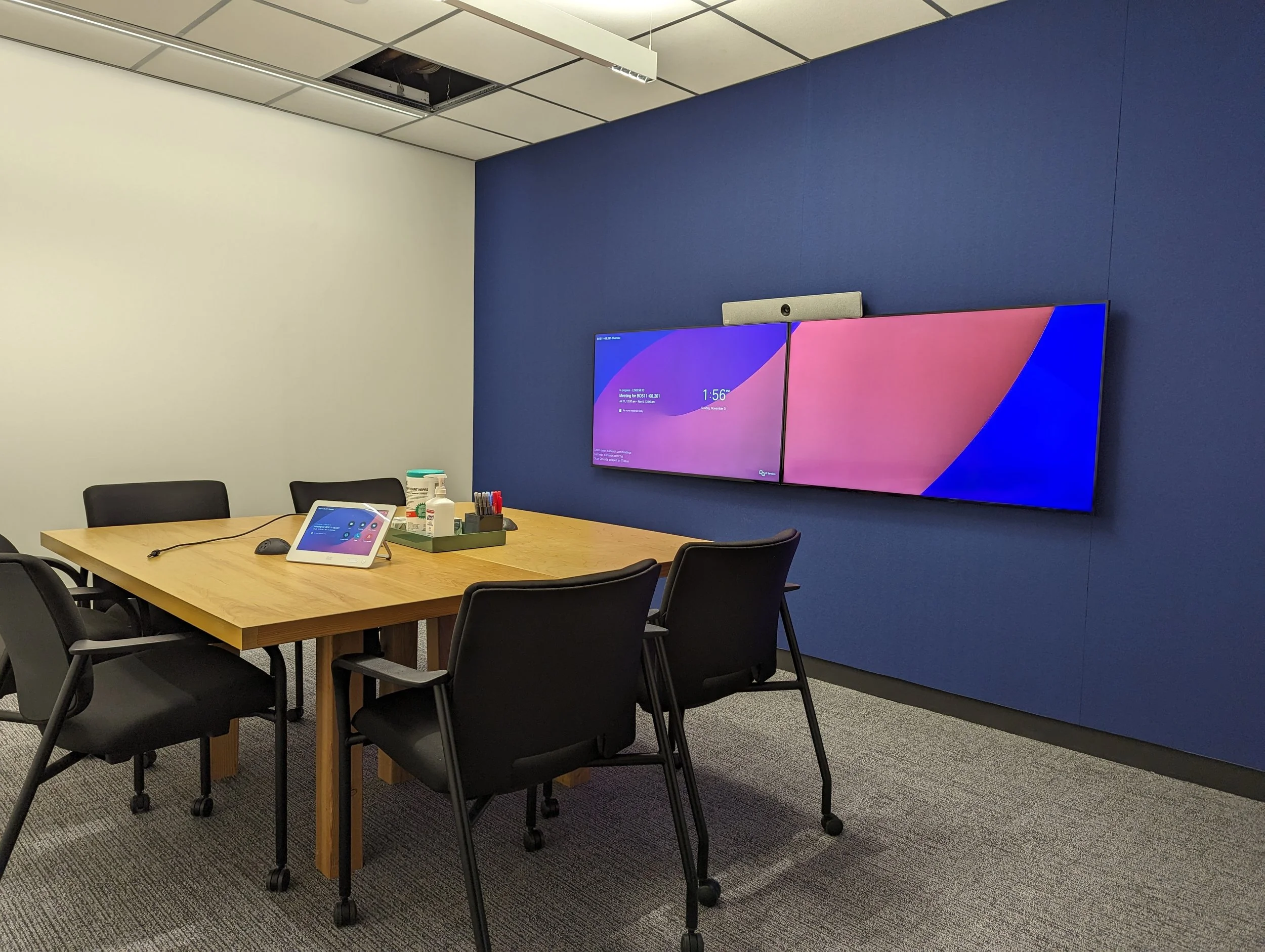 Meeting room with a wooden table, six black chairs, a tablet, a webcam, and cleaning supplies. Mounted on a blue wall is a large dual-screen display. The room has a white and blue wall and a carpeted floor, with a missing ceiling panel.