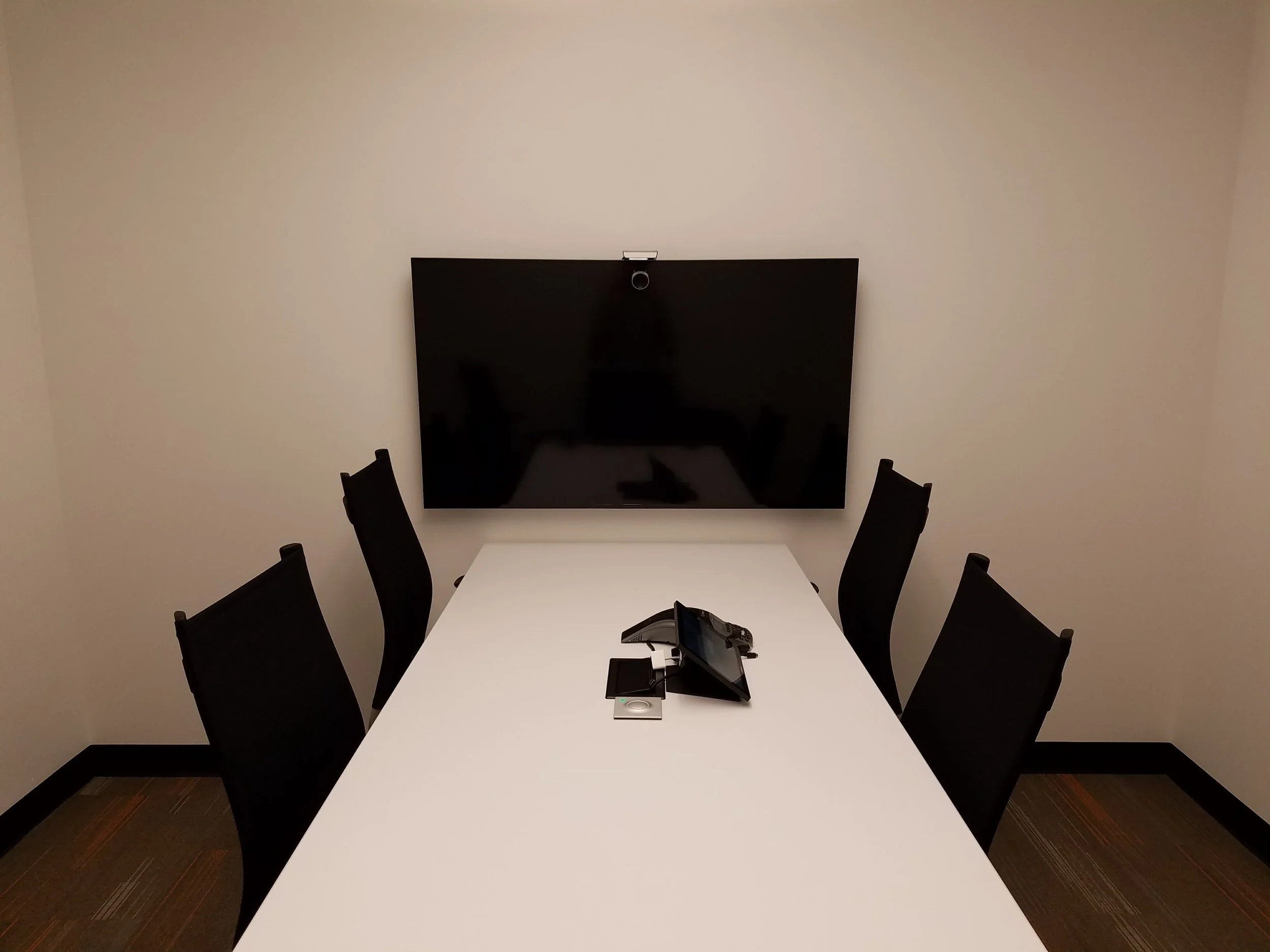 Conference room with a white table, five black chairs, a large wall-mounted TV, and a conference phone on the table.
