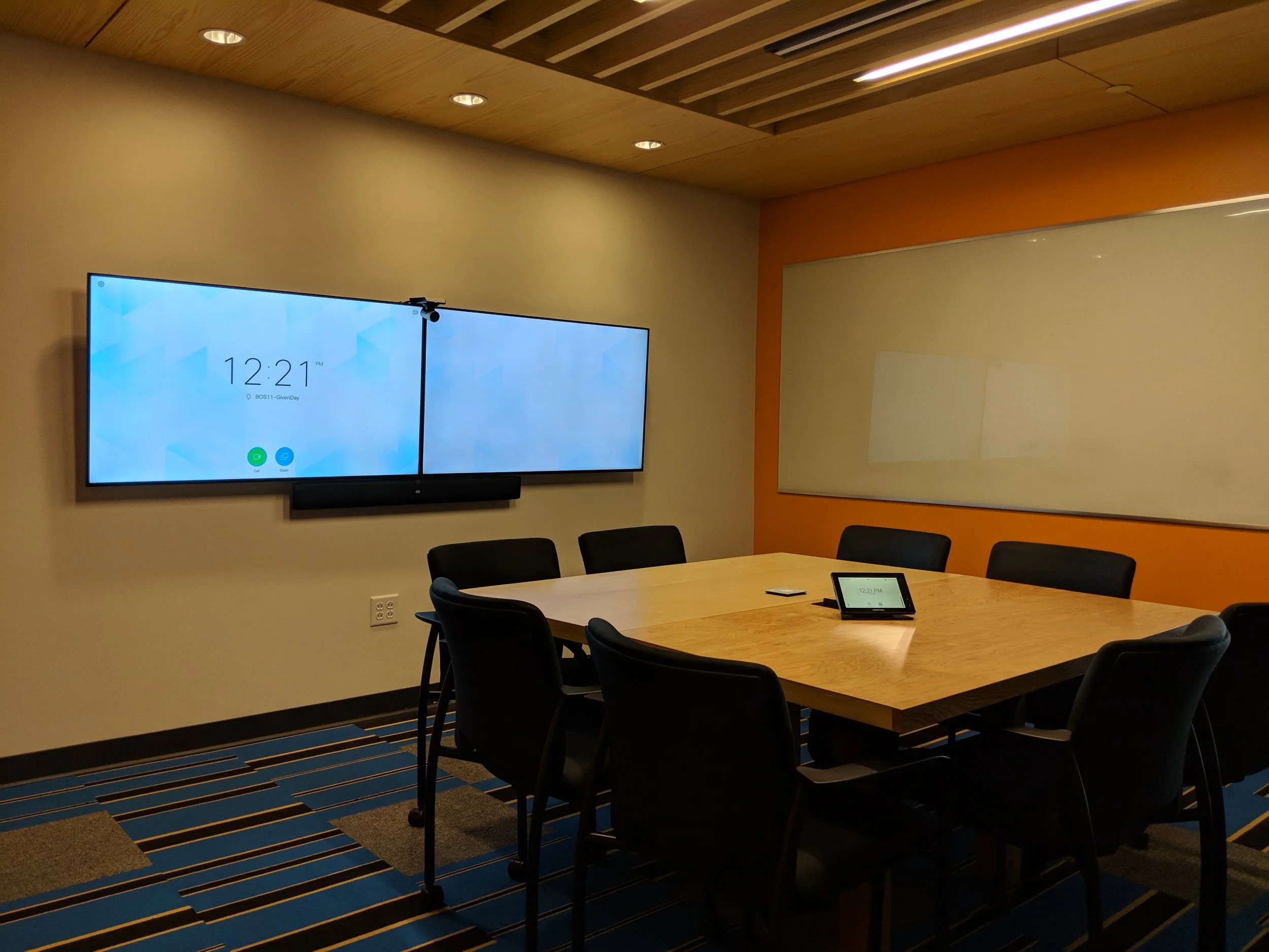 Empty conference room with a wooden table, six black chairs, a large dual-screen monitor on the wall displaying the time, and a tablet on the table.