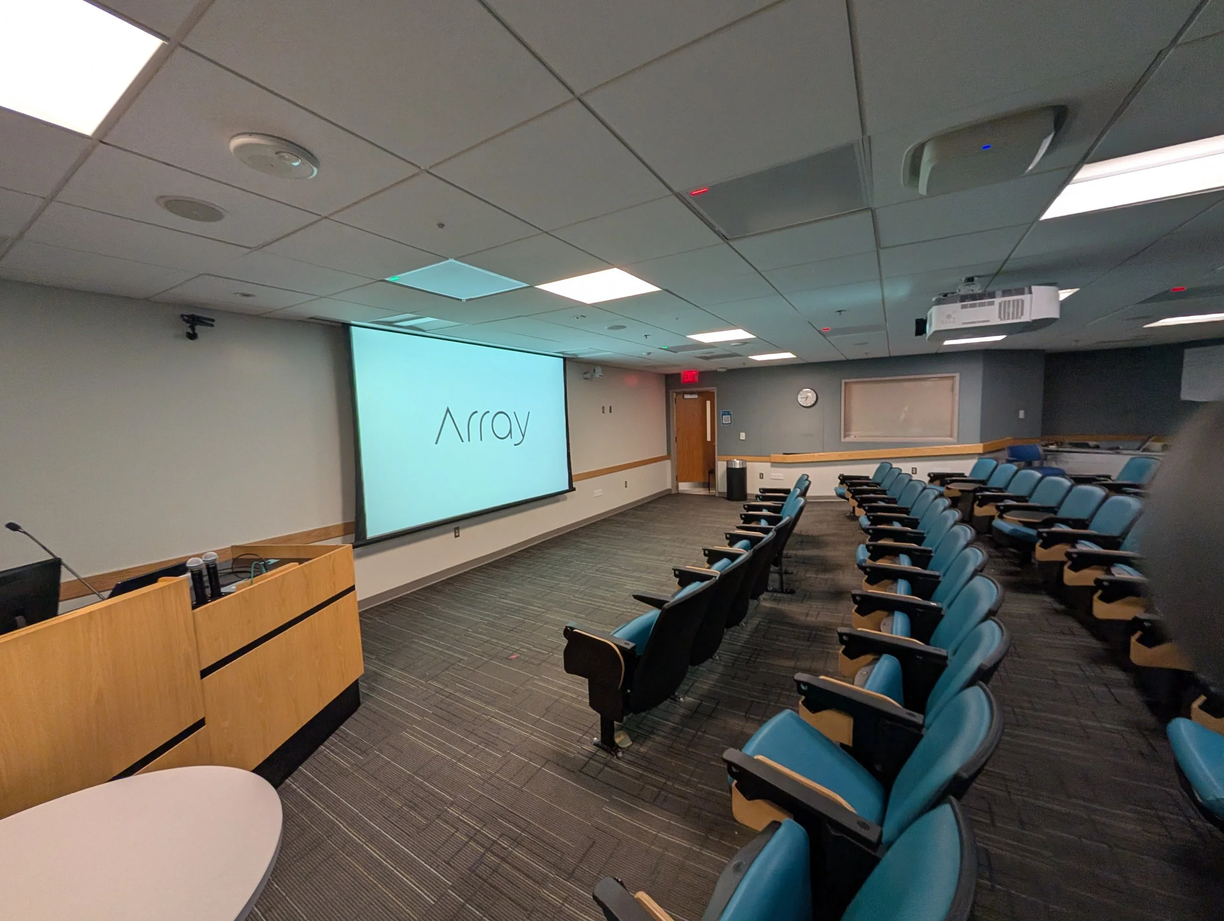 Empty classroom or lecture hall with rows of blue cushioned seats, a large screen displaying the word 'Array', and a wooden podium at the front.