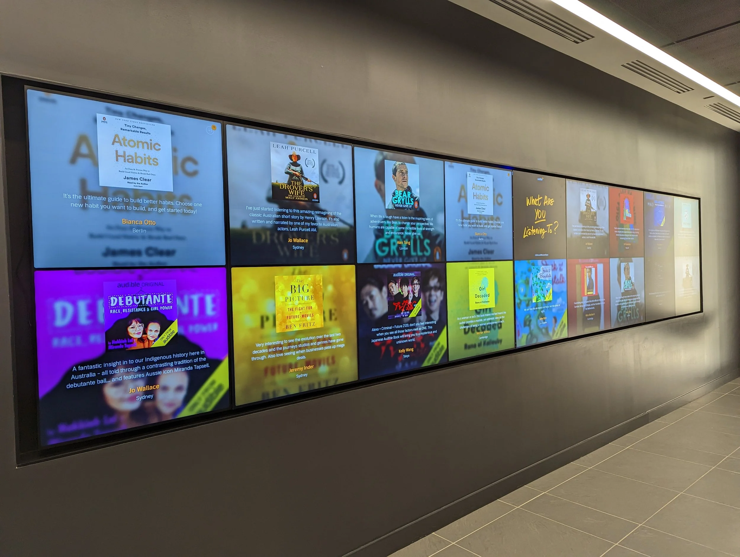 A large digital display wall in an indoor public space showing various colorful posters, book covers, and descriptions, including titles about habits, books, and audiobooks, with a tiled floor and ceiling air vents visible.