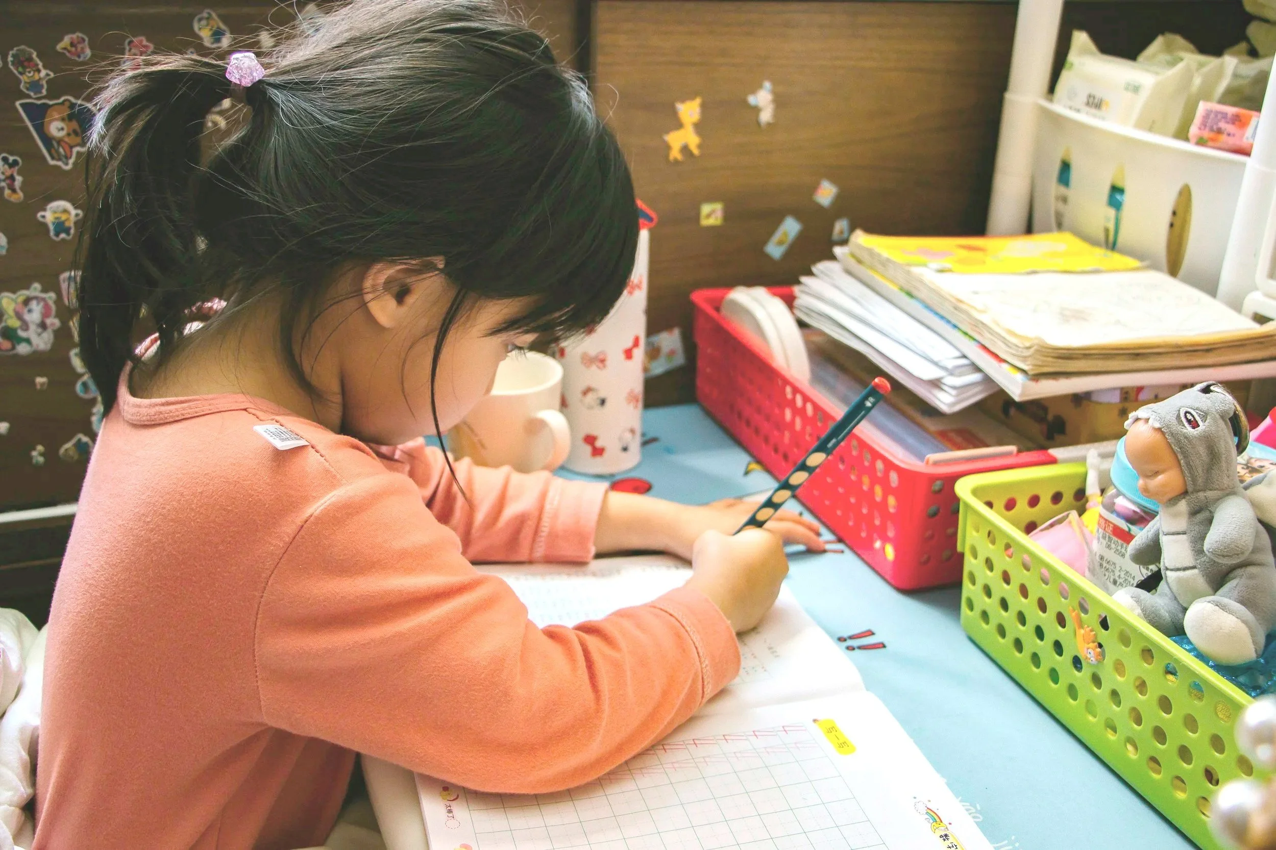 A young girl doing homework at a desk, surrounded by school supplies, stuffed animals, and notebooks.