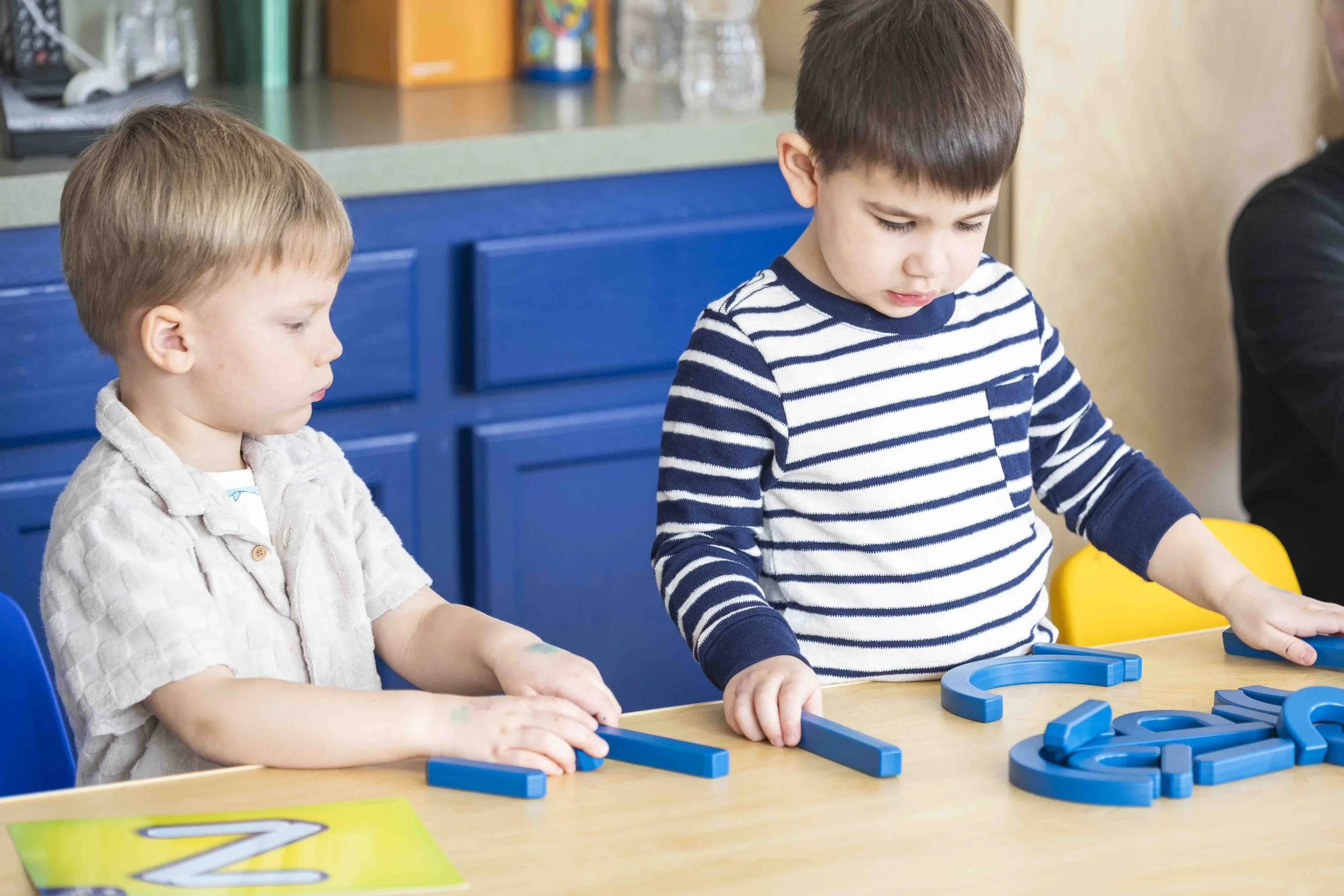 Two young boys seated at a table playing with blue plastic educational toys, with a blue cabinet and books in the background.
