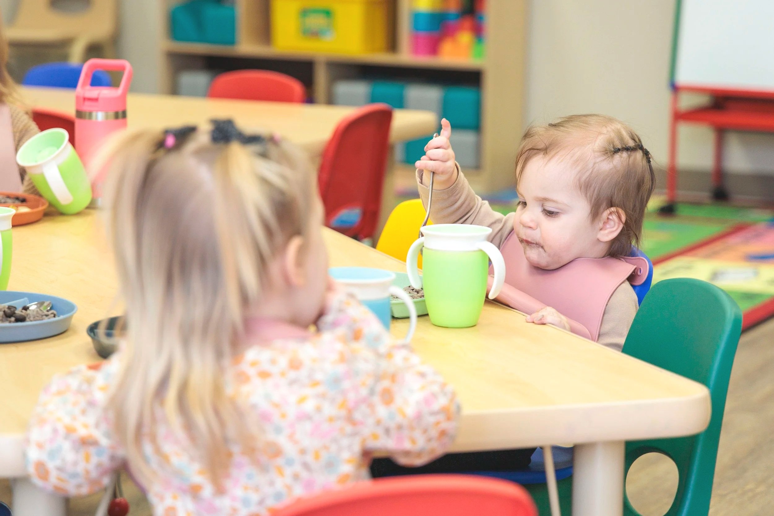 Children eating at a colorful classroom table with cups and bowls.