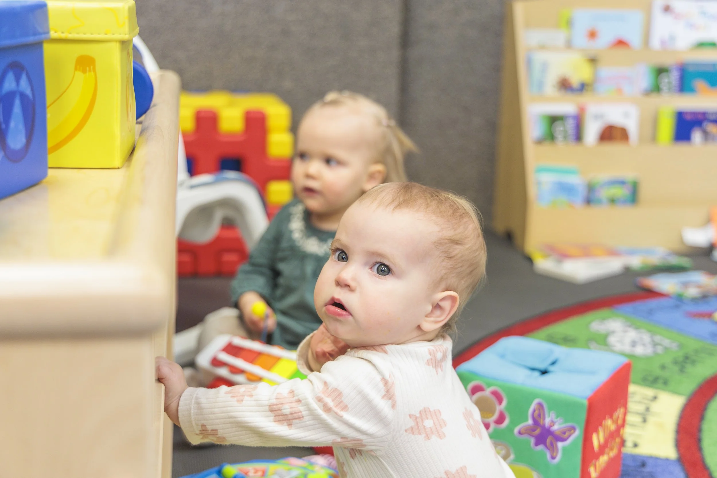 A young child with reddish hair plays on a black and white striped rug, surrounded by toy cars and a pizza slice toy, in a playroom with shelves filled with toys in the background.
