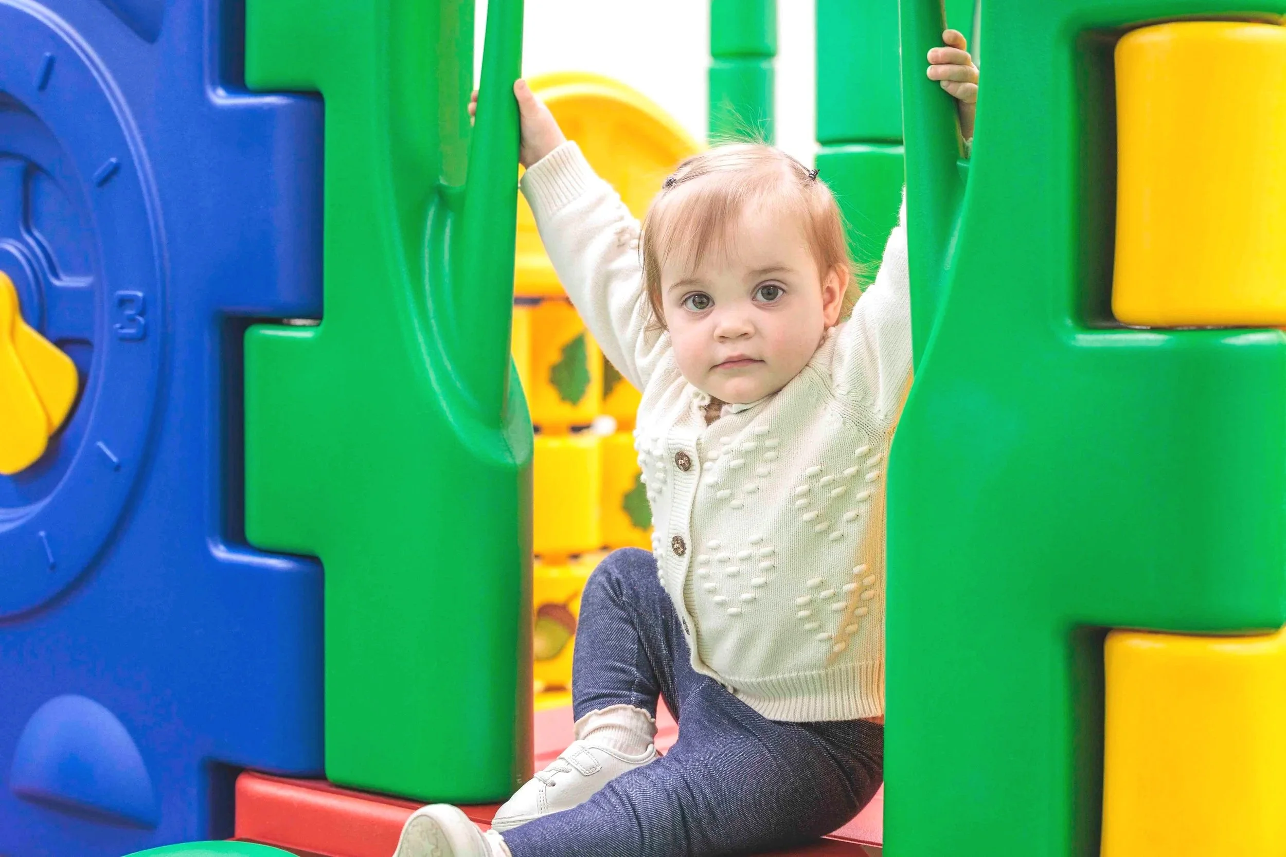 A young child with blonde hair and big eyes playing inside a colorful plastic playground, holding onto green bars and sitting on a red surface with a yellow and blue structure in the background.