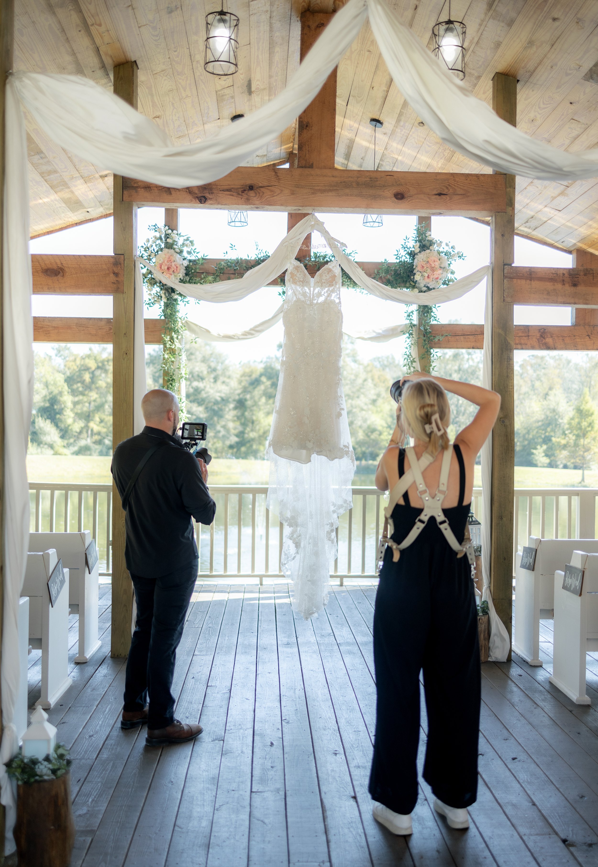 A wedding dress hanging in a decorated outdoor wooden pavilion, with two photographers capturing it.