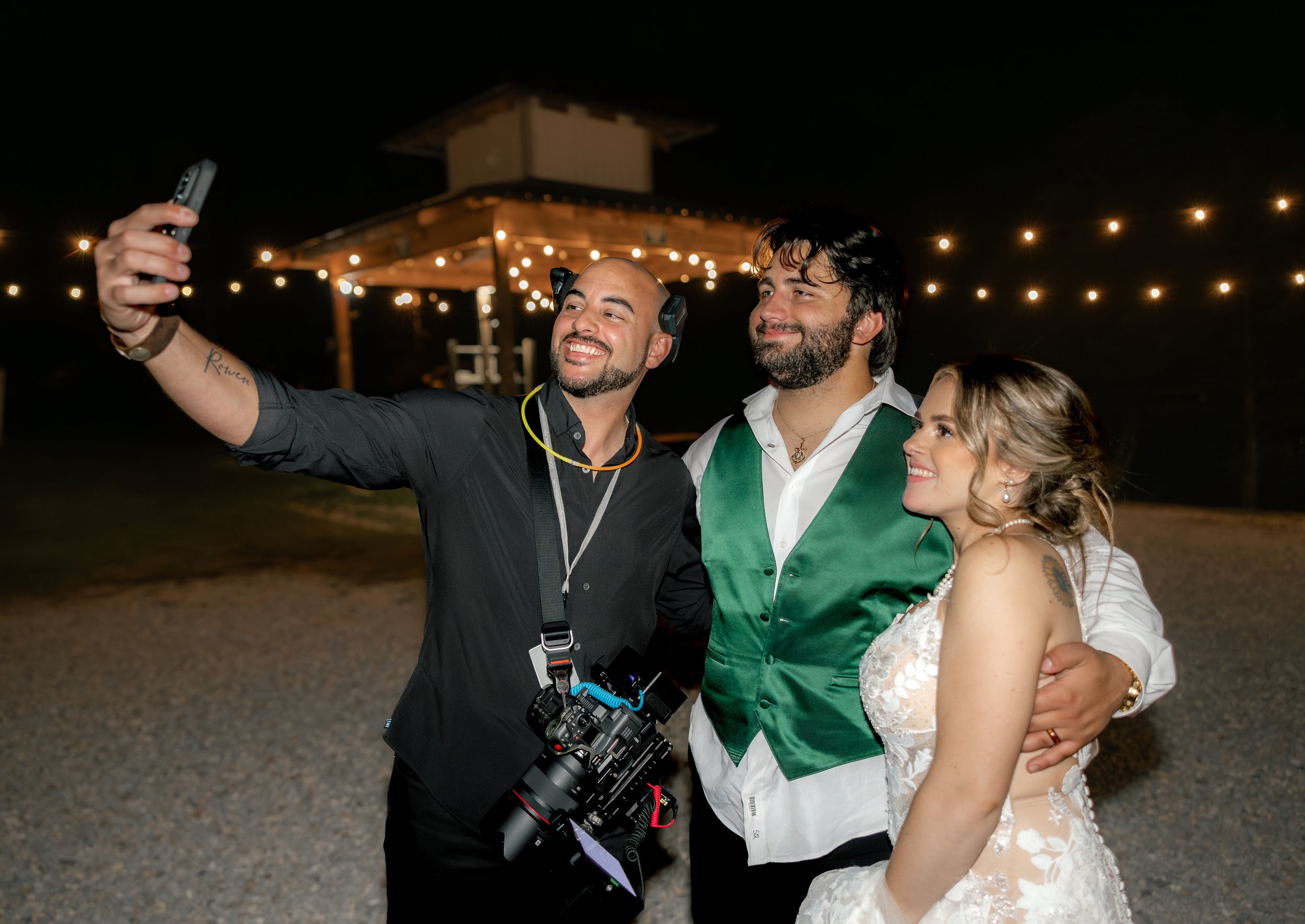 A man taking a selfie with a newlywed couple at an outdoor wedding, with string lights in the background.