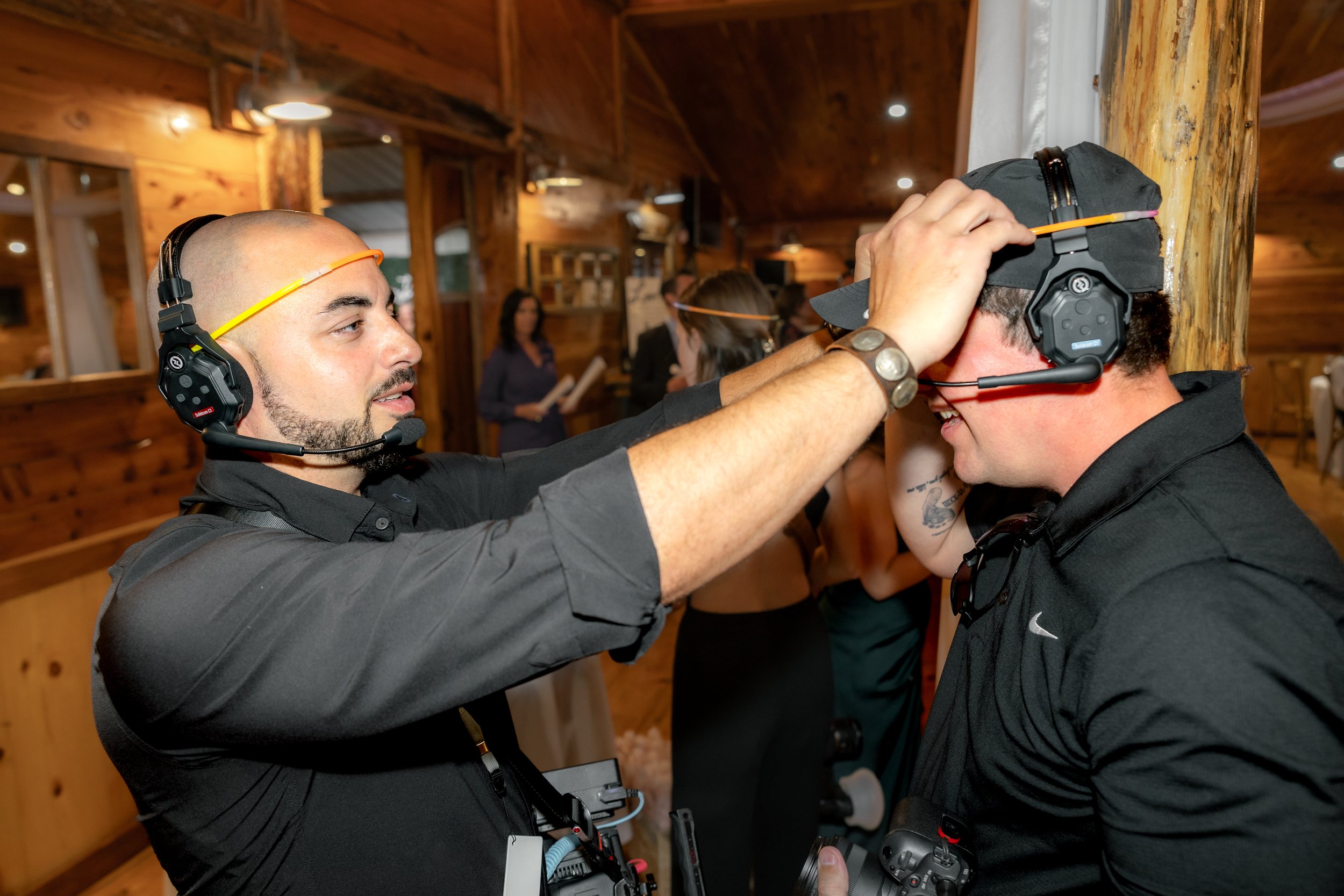 Two people wearing headsets in an indoor event setting, one adjusting the other's headset, with cameras around their necks.