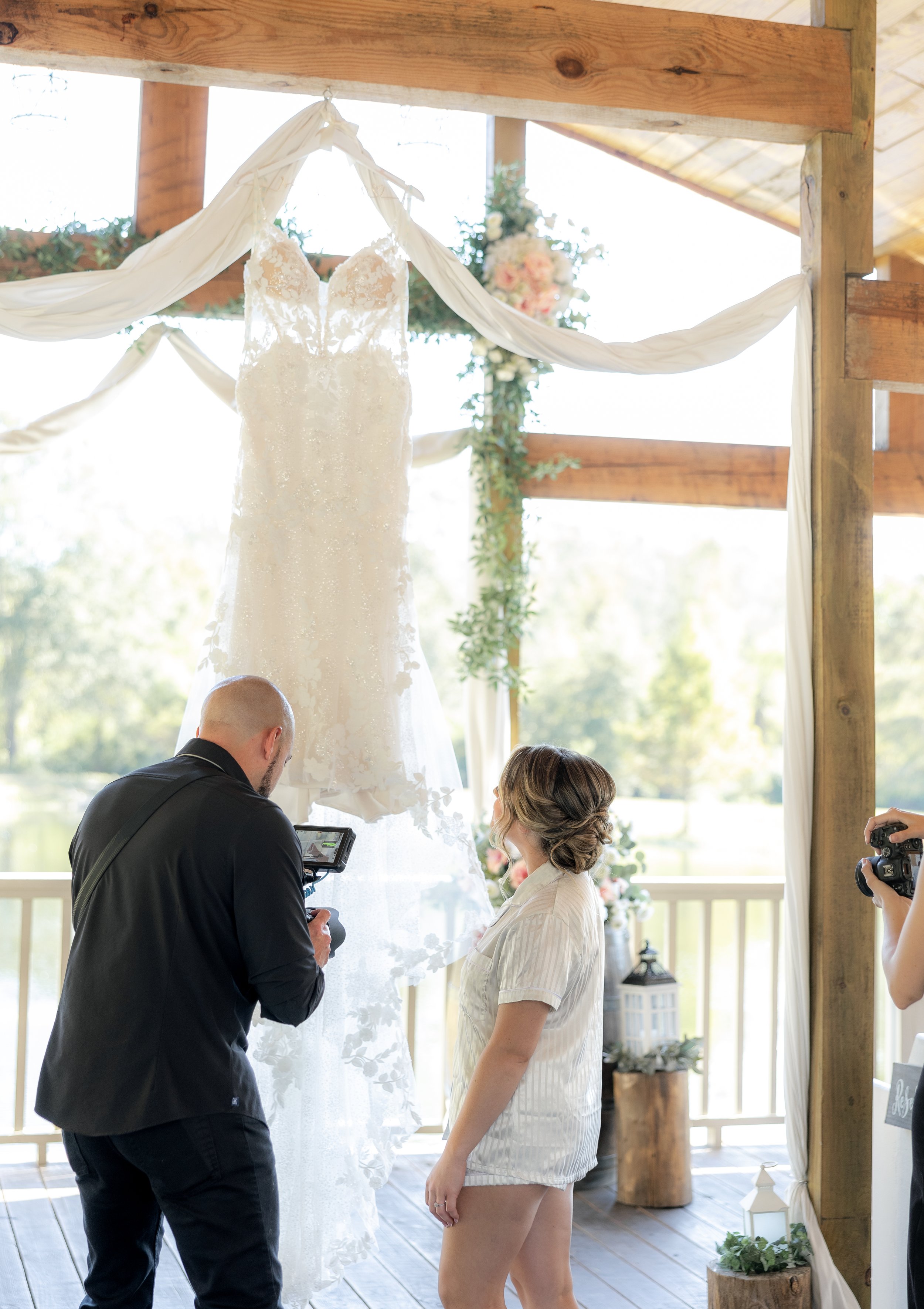 A wedding dress hanging on an archway with two photographers and a woman observing it, set in a wooden venue with decorations.
