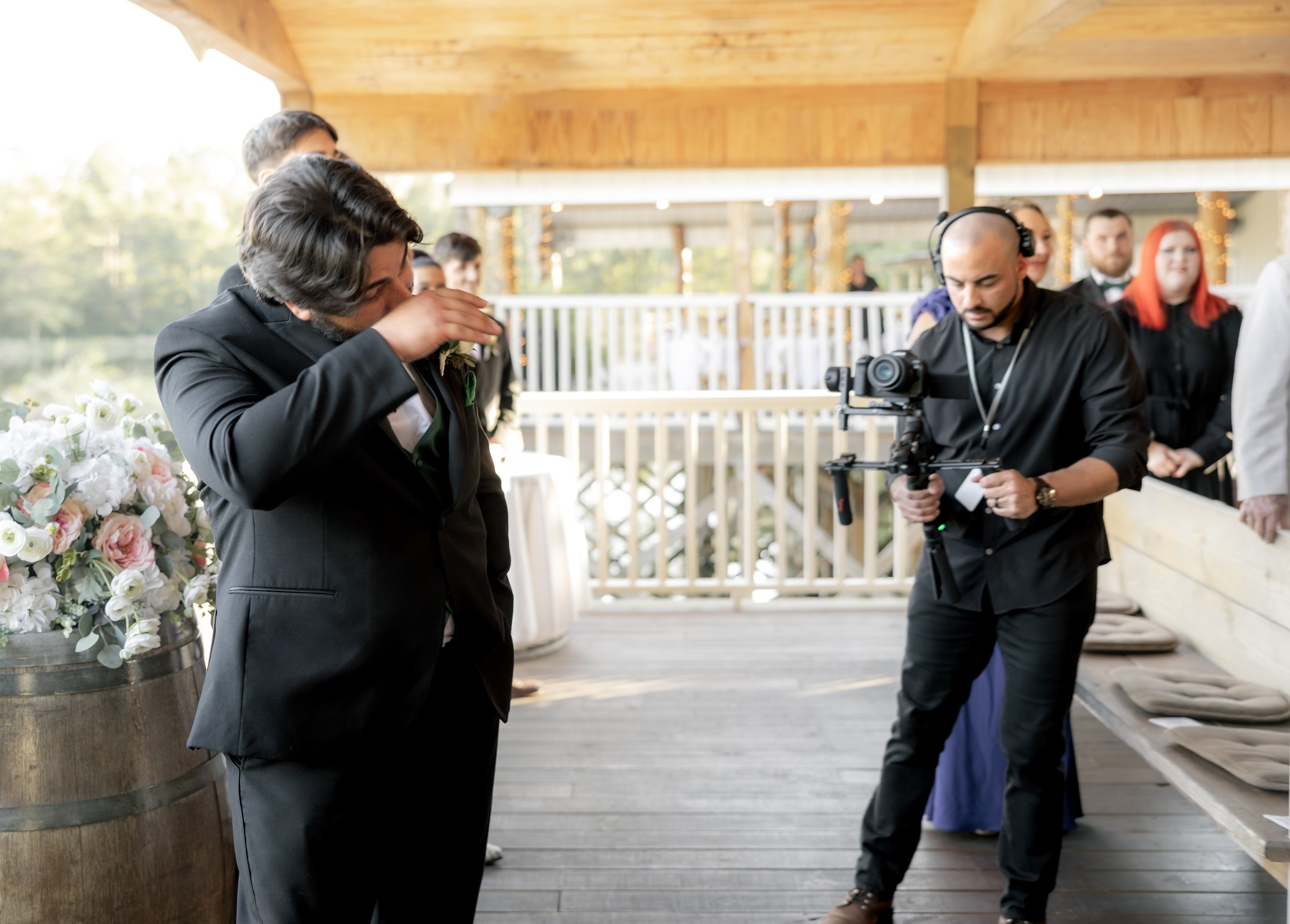 A man in a suit wipes his face at an outdoor event, standing next to a floral arrangement on a wooden barrel. A videographer with a camera on a stabilizer records the scene, and several people stand in the background on a deck with wooden railings.