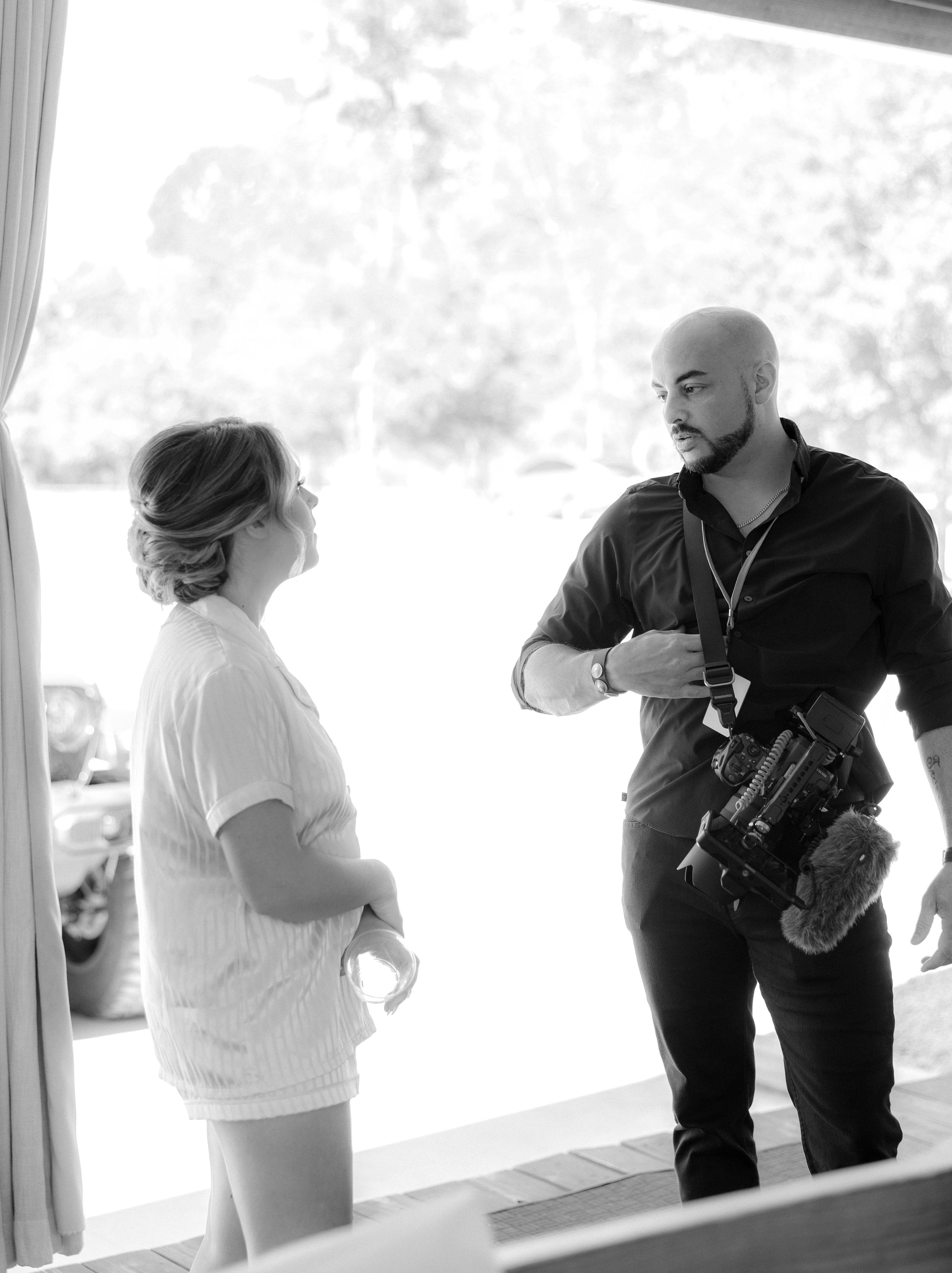 Black and white photo of a woman in casual attire holding a drink, talking to a man with a camera hanging around his neck, engaged in conversation outside a building.