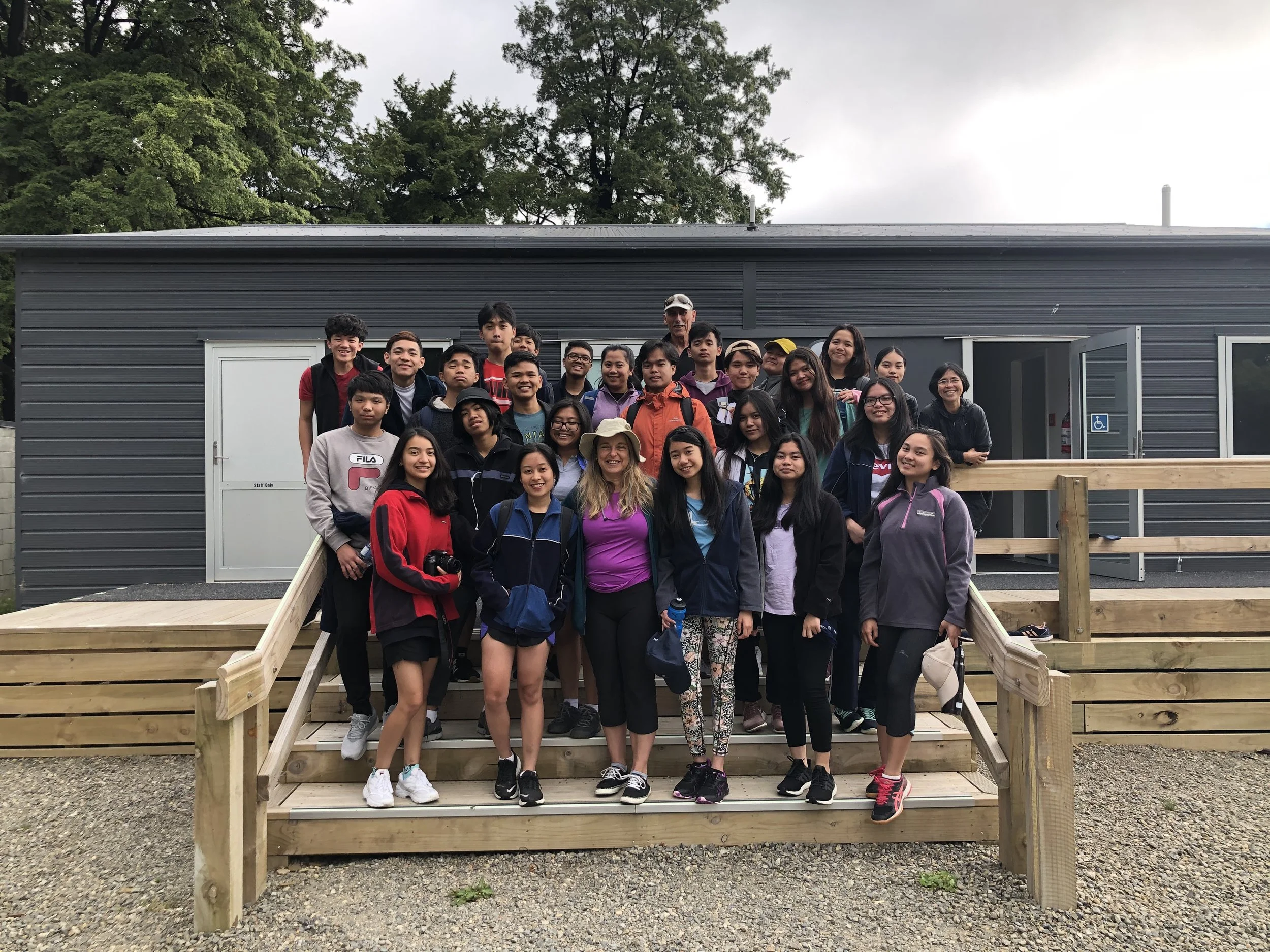 A large group of diverse young students and two adults pose for a photo on the wooden steps in front of a gray building outdoors.