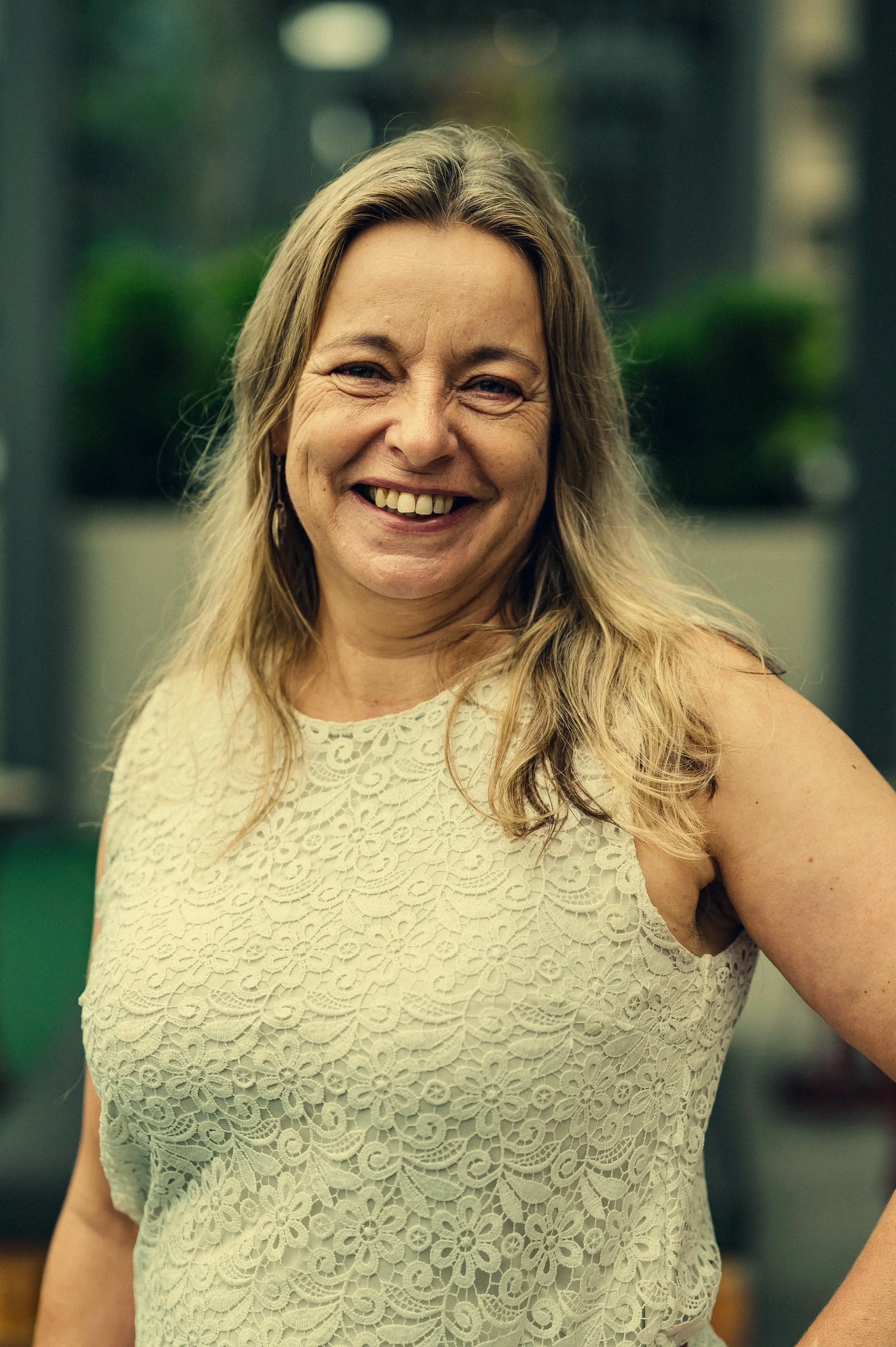 A smiling woman with long blonde hair, wearing a cream-colored lace sleeveless top, standing outdoors with blurred greenery in the background.