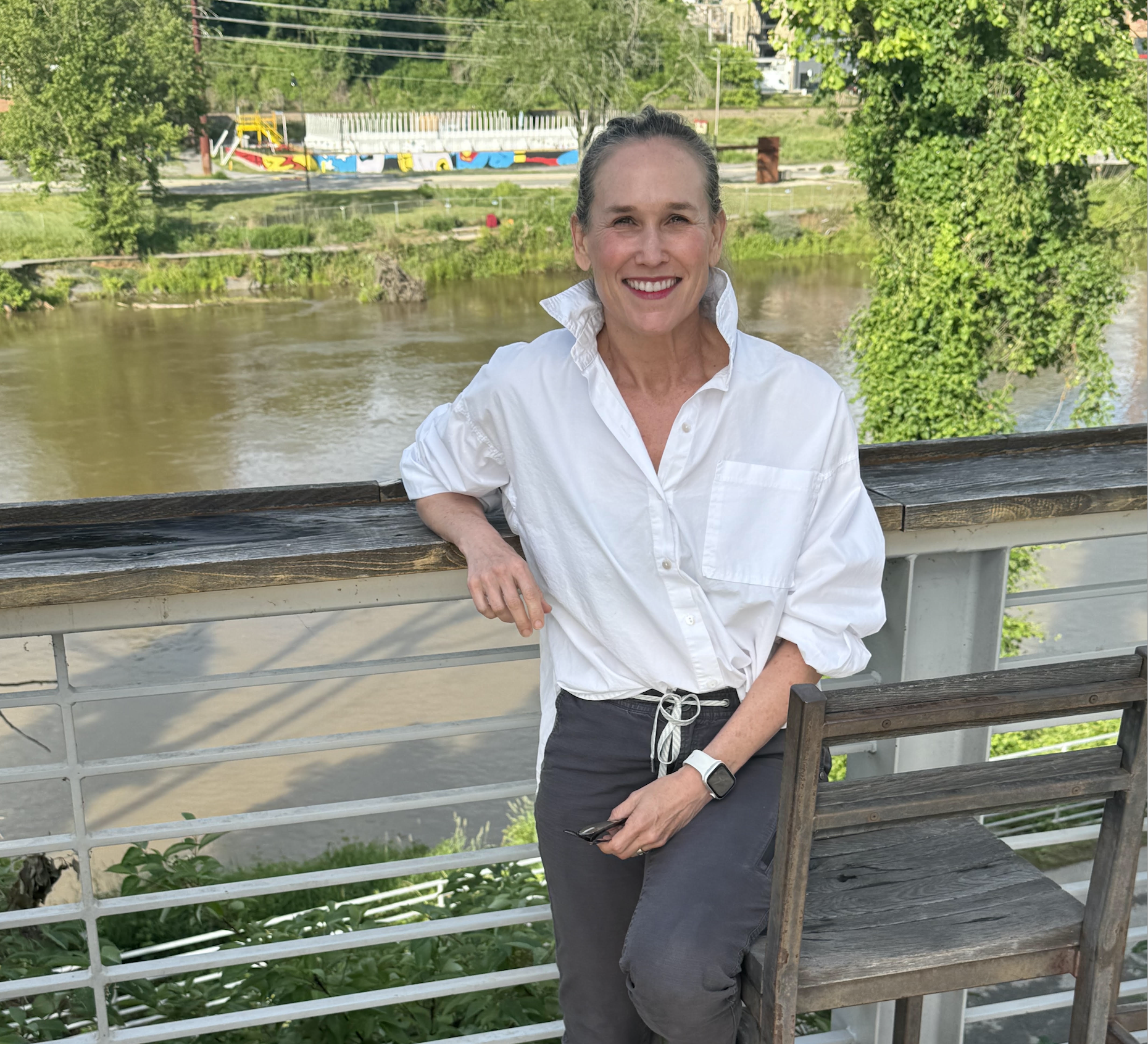 A smiling woman with long blonde hair, wearing a black North Face jacket and jeans, standing by a river with snow-covered trees in the background.