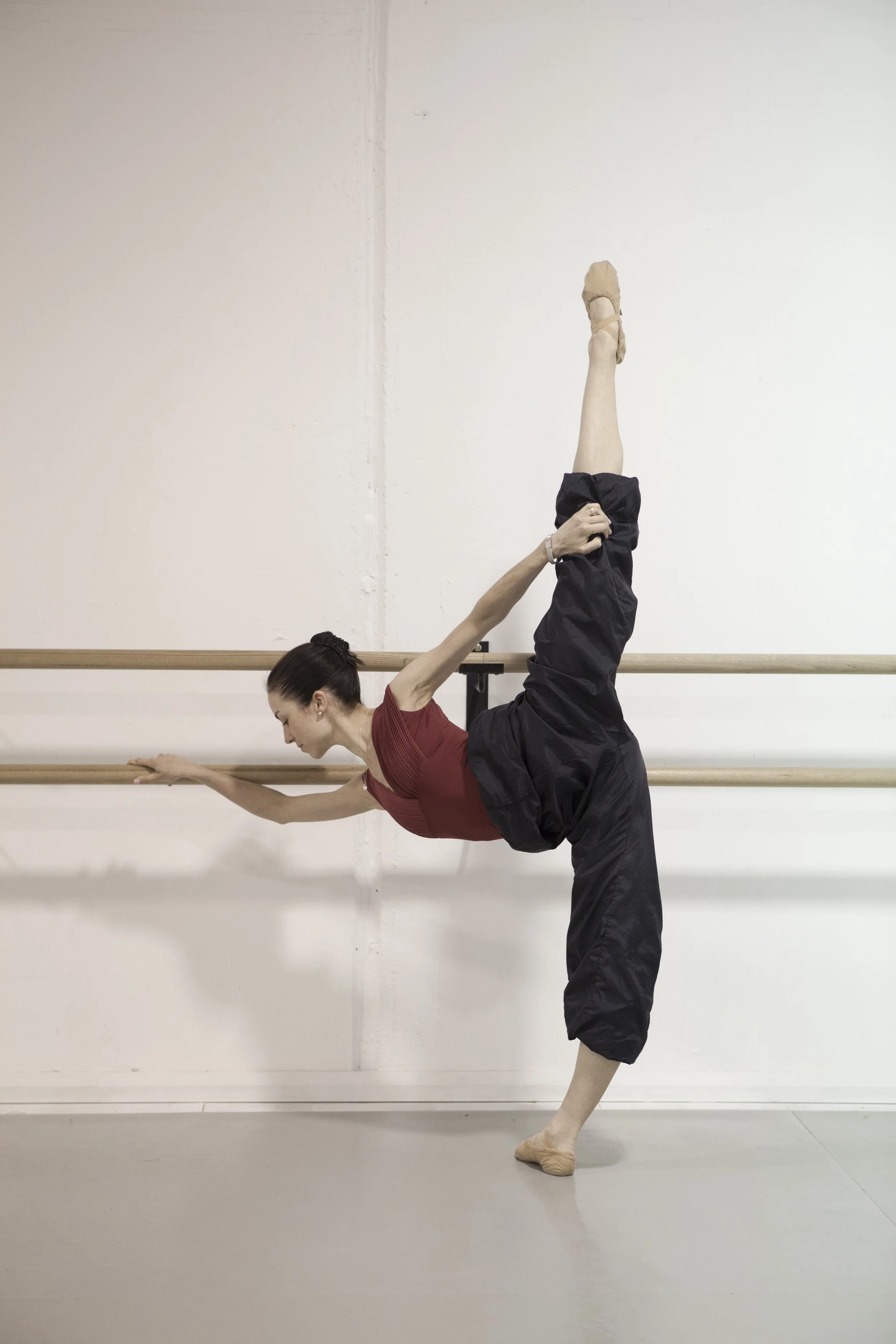 A woman in a dance studio performing a standing split stretch while holding onto a ballet barre.