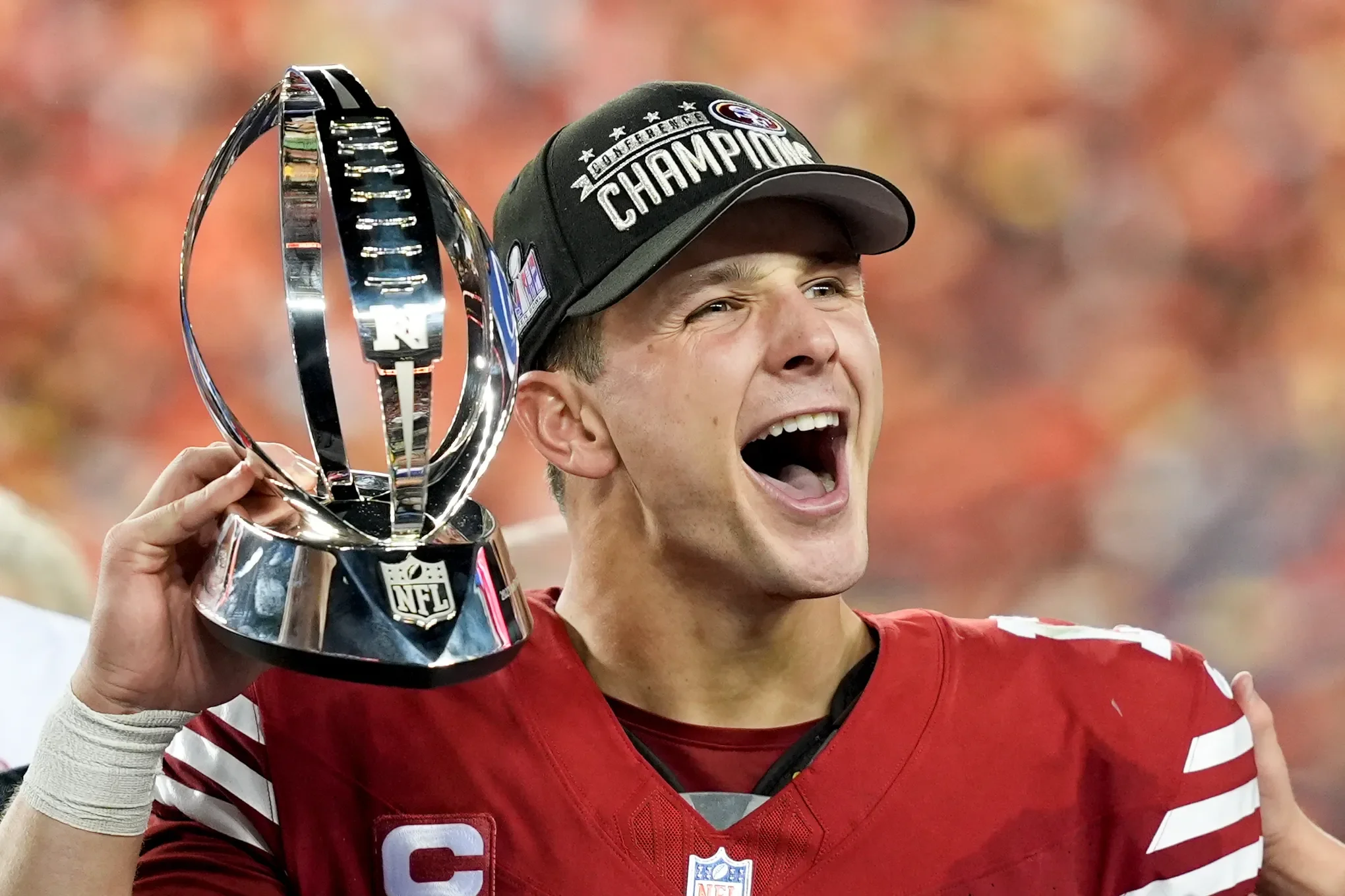 A football player celebrating with a trophy, wearing a cap that reads 'Conference Champions', a red jersey with white stripes, and smiling widely.