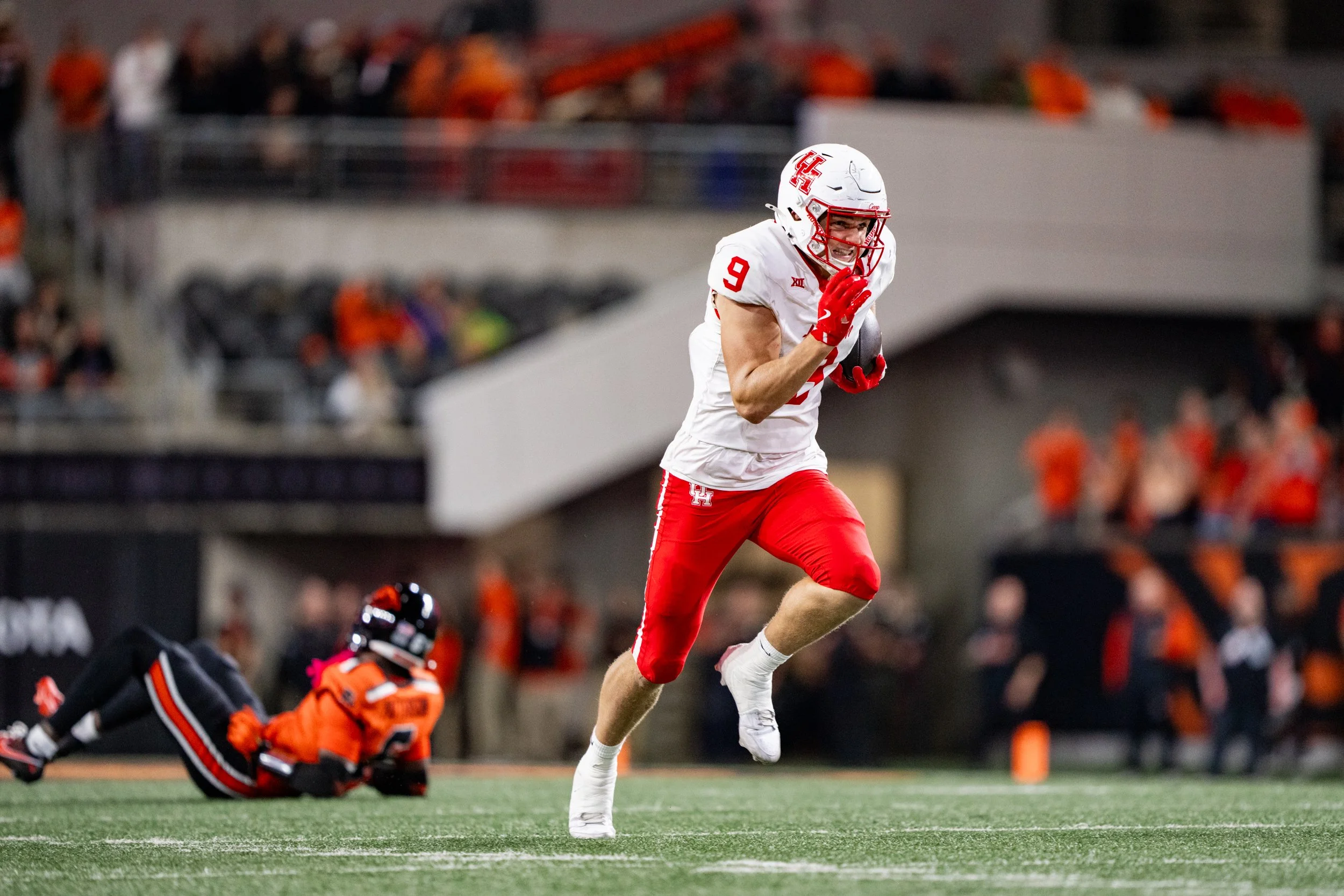 A football player in a white and red uniform running on the field holding a football, with a player in an orange and black uniform lying on the ground in the background.