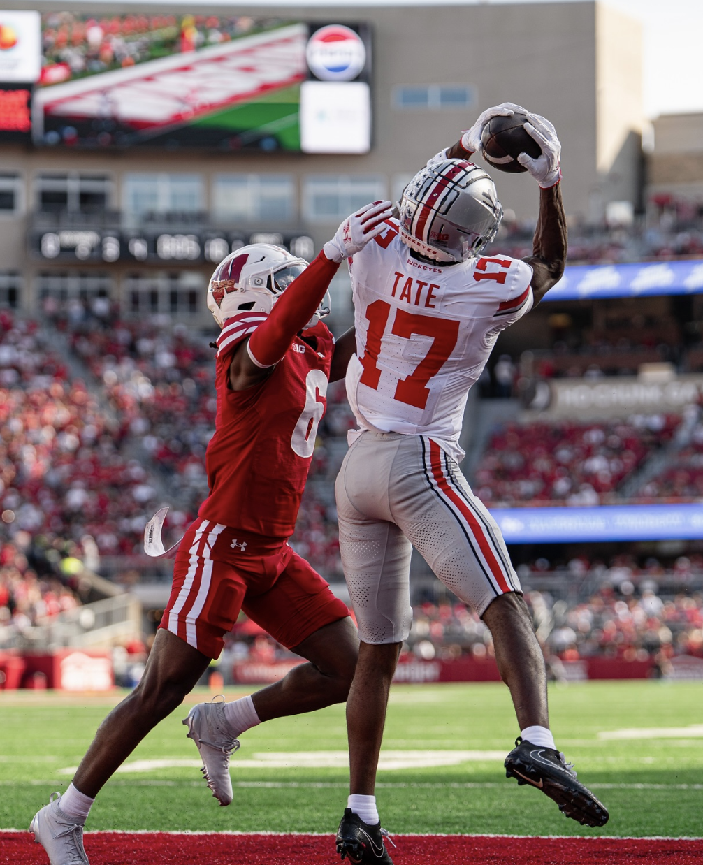 A football player in a white jersey with number 17 and the name TATE catches a football in the end zone while an opposing player in a red jersey with number 6 attempts to defend him during a game.