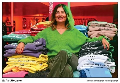 A woman smiling while sitting between stacks of colorful sweatshirts inside a store.