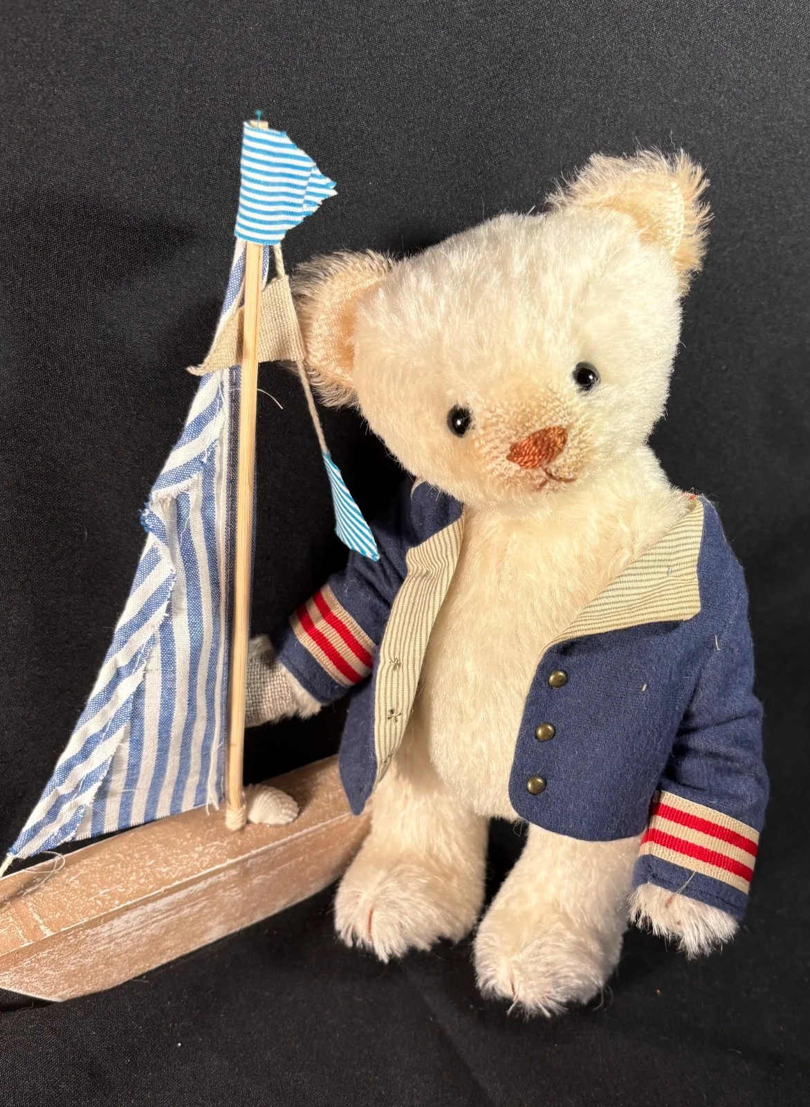 Teddybär Lars in Matrosenjacke sitzend neben einem Holzsegelboot, frontal