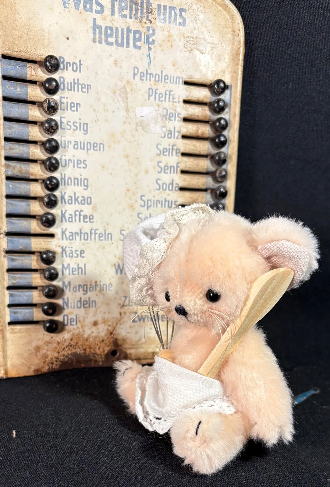 Bär mit weißer Spitzenhaube liegt vor einer alten Einkaufstafel, mit Schneebesen und Holzlöffel in der Stoffschürze