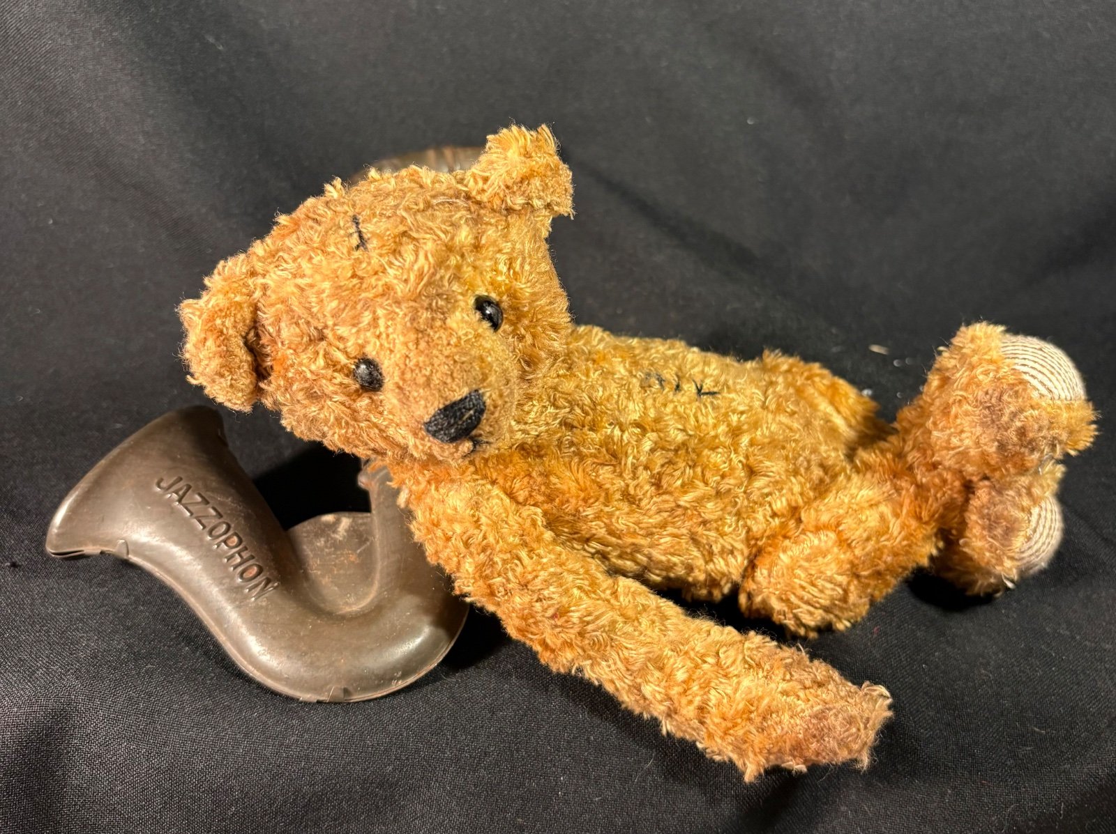 Teddybär Emil liegend mit seitlich geneigtem Kopf, daneben ein altes Blechspielzeug in Saxophonform