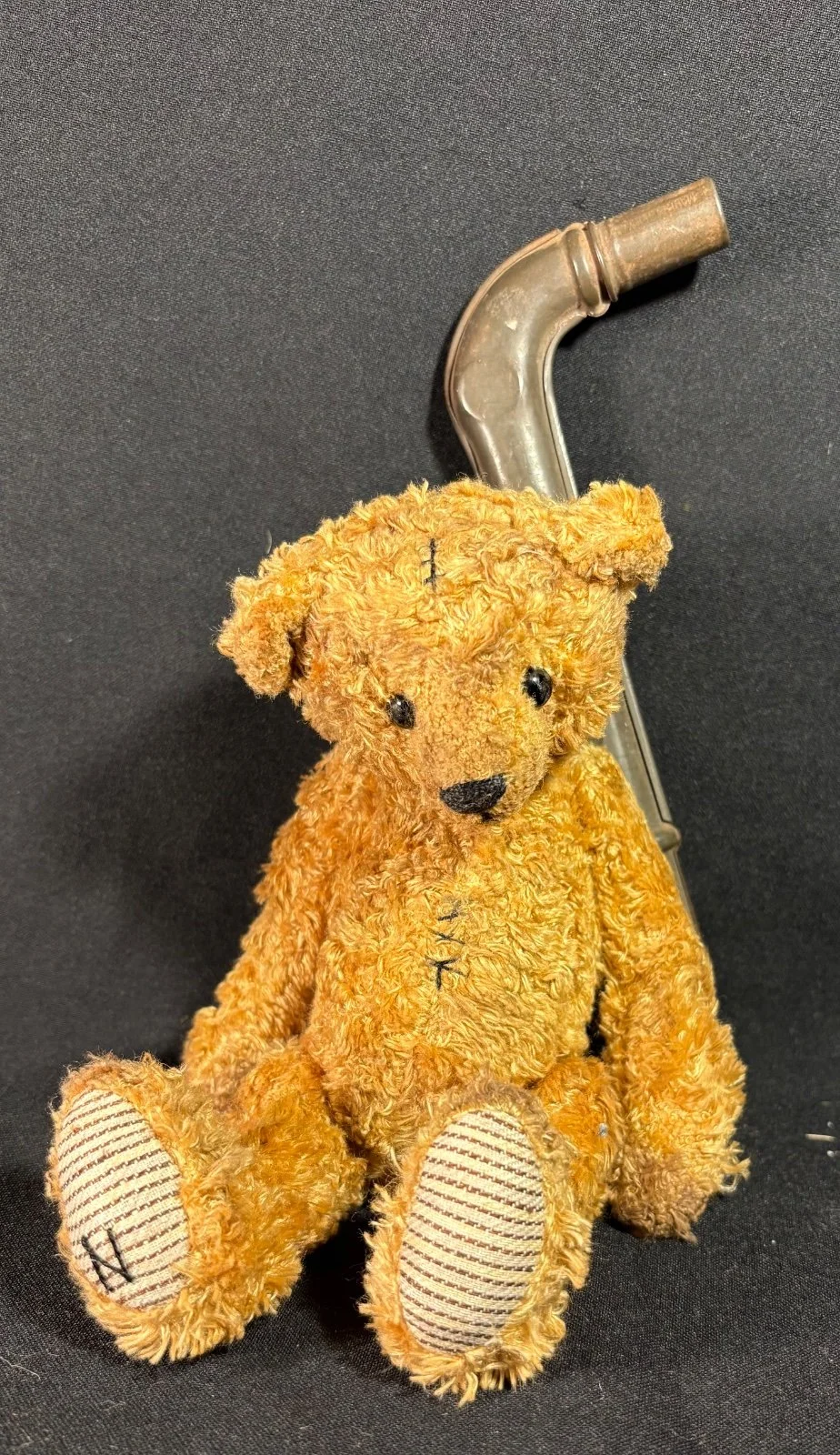 Teddybär Emil sitzend von vorn, Fußsohlen mit gestreiftem Stoff sichtbar, im Hintergrund das Blechspielzeug