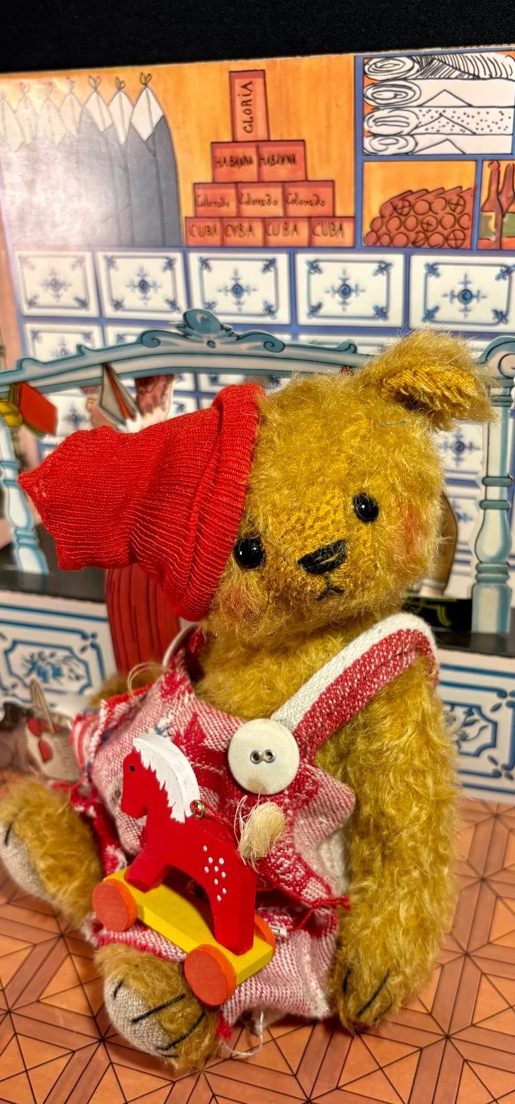 Teddybär Pelle mit roter Zipfelmütze und Latzhose, sitzend vor Holzkulisse mit Ziehpferd