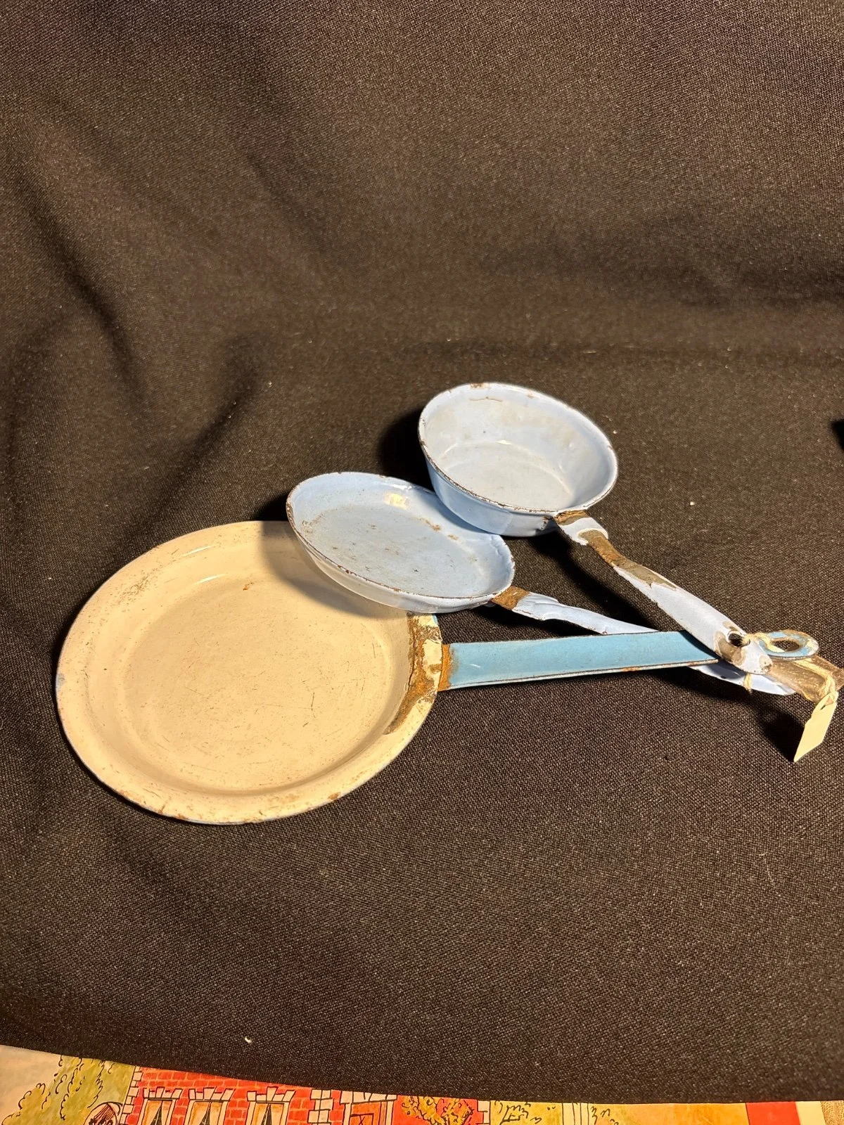 Drei kleine Puppengeschirr-Pfannen mit Stielen, eine creme- und zwei blaufarben, auf dunklem Stoff
