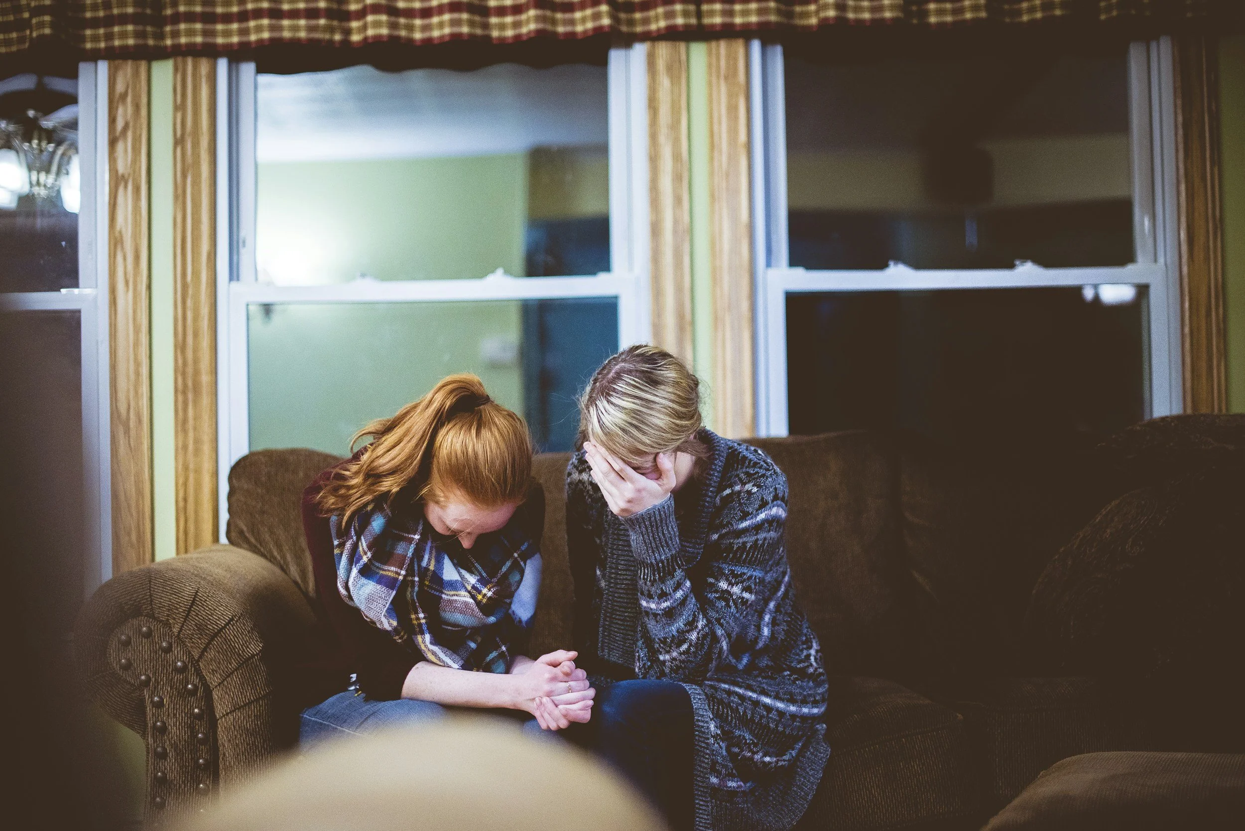 Two women sitting on a sofa, one with red hair and the other with blonde hair, sharing a moment of laughter and comfort, in a living room.