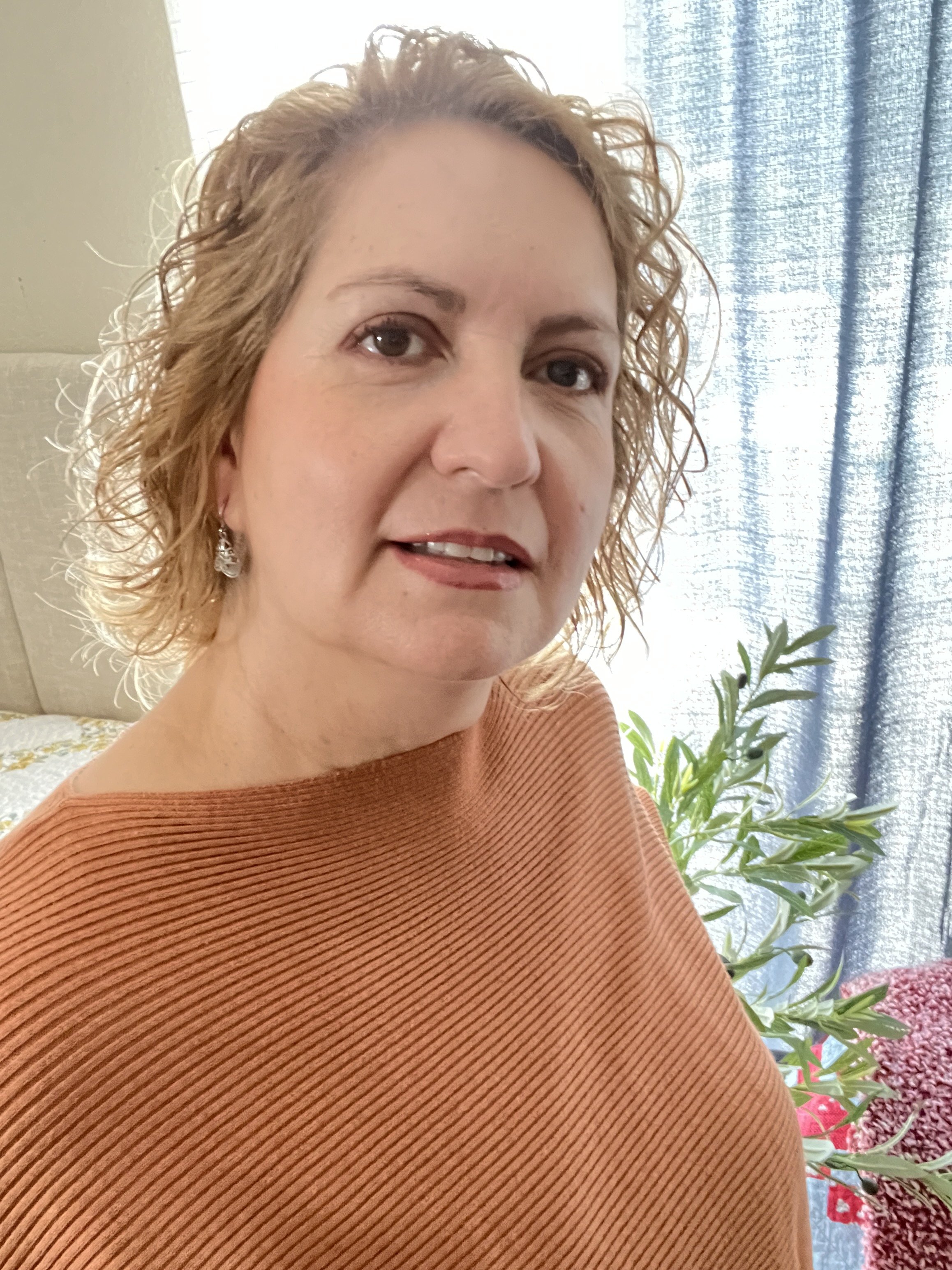 A woman with curly blond hair and light skin, wearing a rust-colored, ribbed top, standing indoors near a window with blue curtains and green plants in the background.